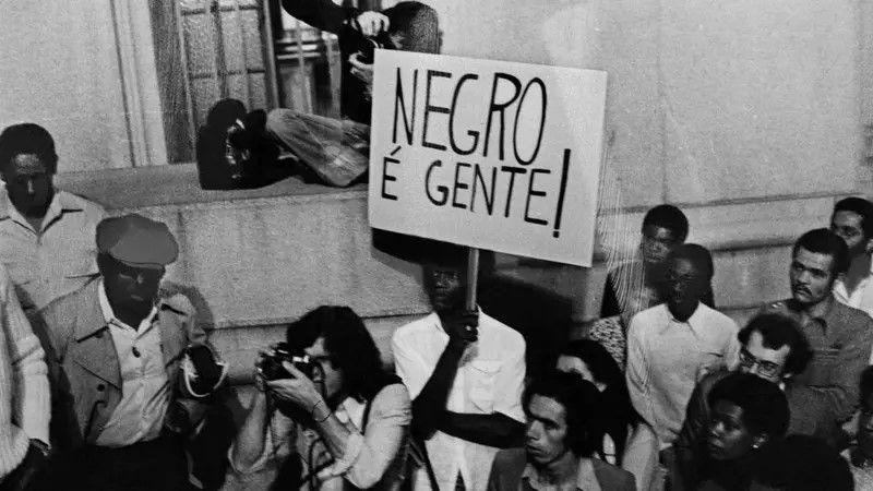 Homens negros em uma manifestação. Um deles segura uma placa escrito: Negro é gente!