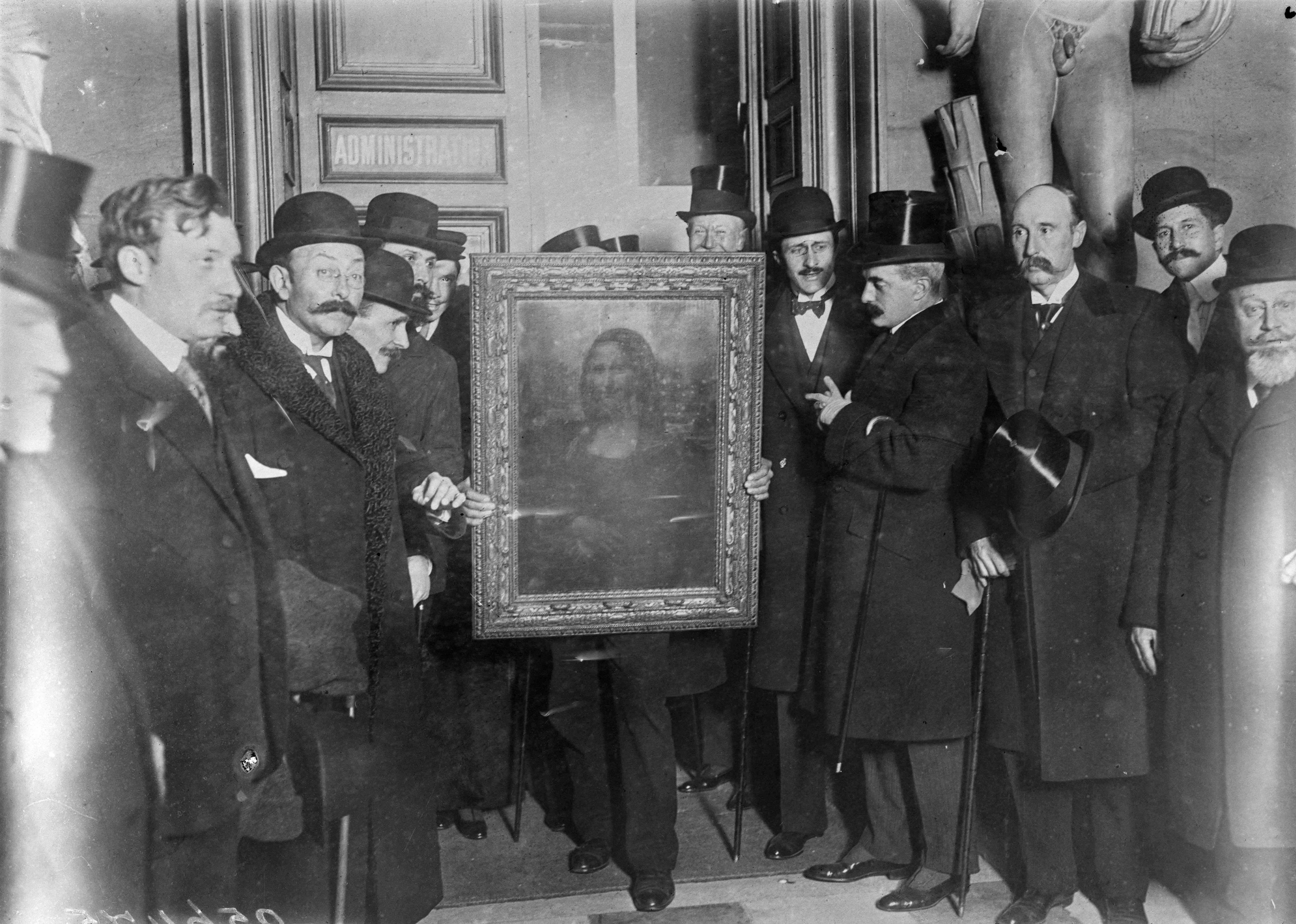 Imagen en blanco y negro de 1914 de un grupo de hombres vestidos con elegantes abrigos, trajes, corbatas y sombreros negros. En el centro, una imagen original enmarcada de la Mona Lisa