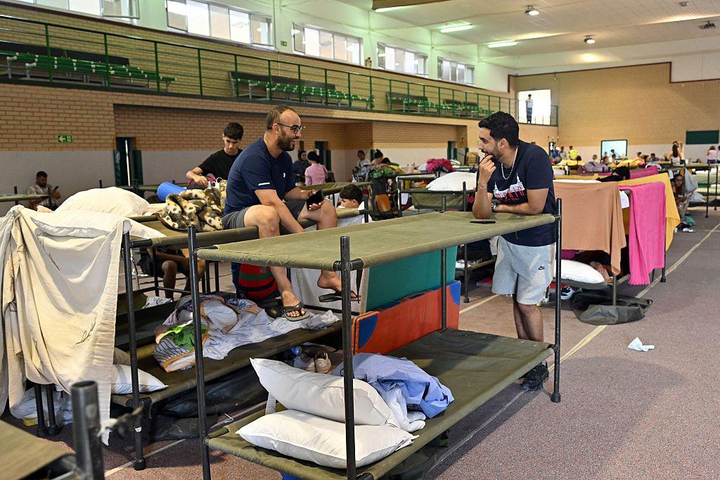 Literas de campaña hechas con tubos y lona de color militar se alienean en un polideportivo. La gente ha puesto en ellas sus almohadas, sábanas o ha colgado sus toallas. En una de ellas, sentado en la parte de arriba, hay un hombre con pantalón corto y camiseta de manga corta que conversa con otro, que está apoyado en otra de las literas y también viste pantalón corto y camiseta de manga corta. Al fondo se ve más gente. 