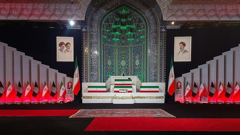 توابيت مغطاة بأعلام إيرانية في باحة مسجد بساط أحمر وأعلام إيران ورايات إسلامية - التابوت الأول من الأعلى فوقه عمامة سوداء في إشارة إلى جثمان علي خامنئي