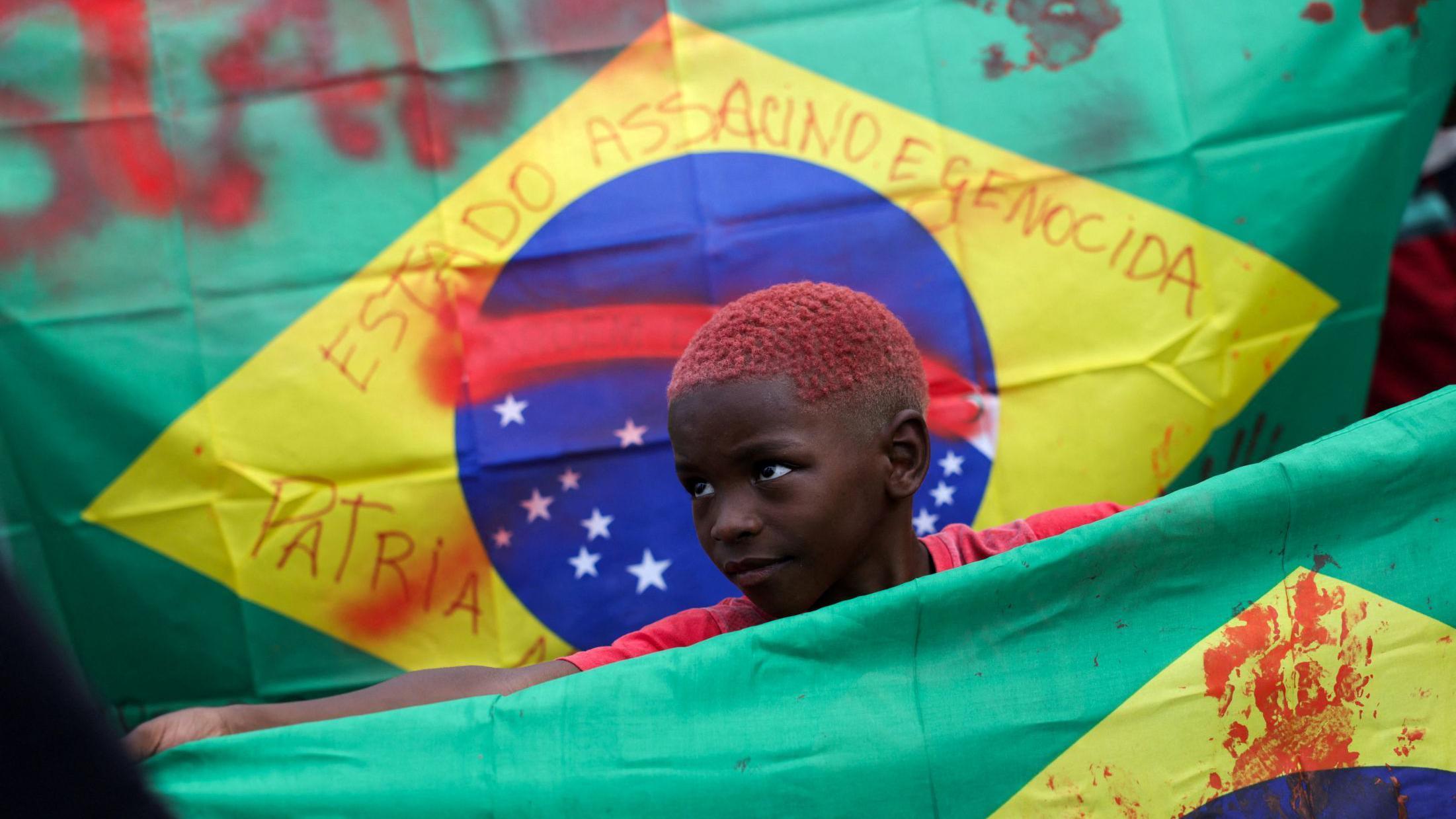 Menino segura bandeira do Brasil manchada com tina vermelha em manifestação.