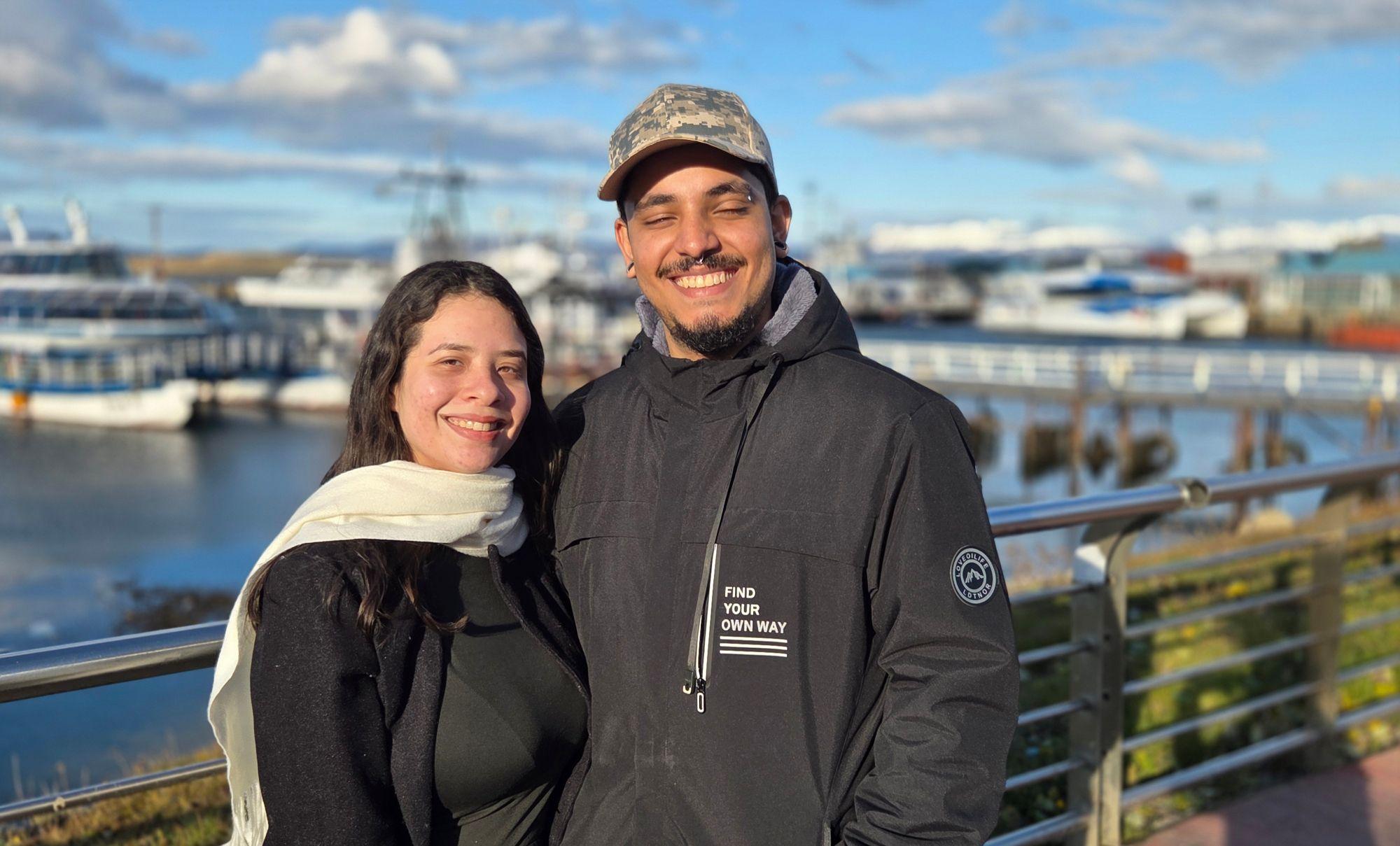 Una pareja sonríe a la cámara con un puerto de fondo