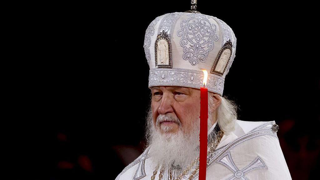El Patriarca de Moscú vestido de blanco y con una vela roja en la mano en una ceremonia.