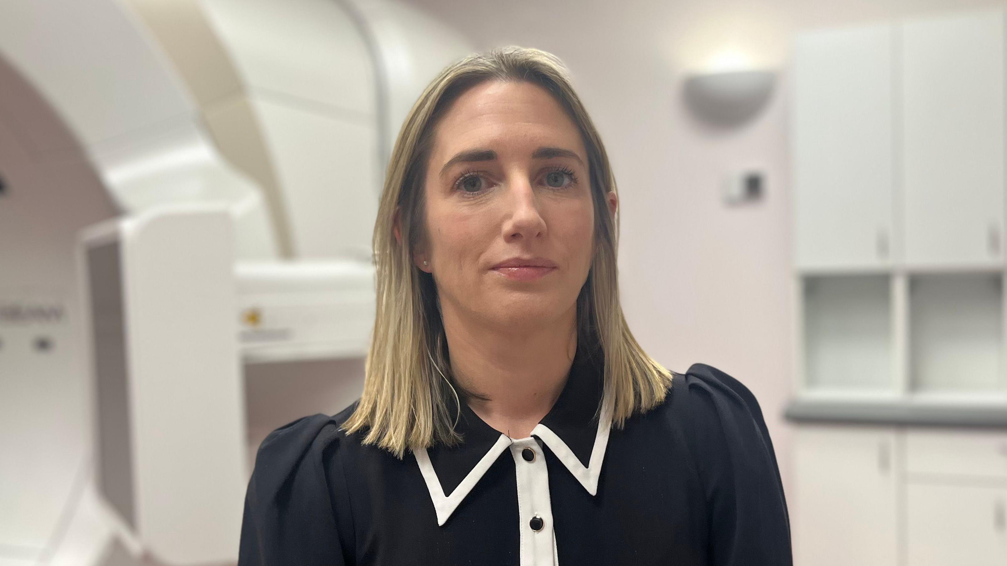 Dr Hanna has shoulder length blonde hair and is wearing a black top with white trim and standing in a hospital room, behind her the edge of a large white medical scanner can be seen.