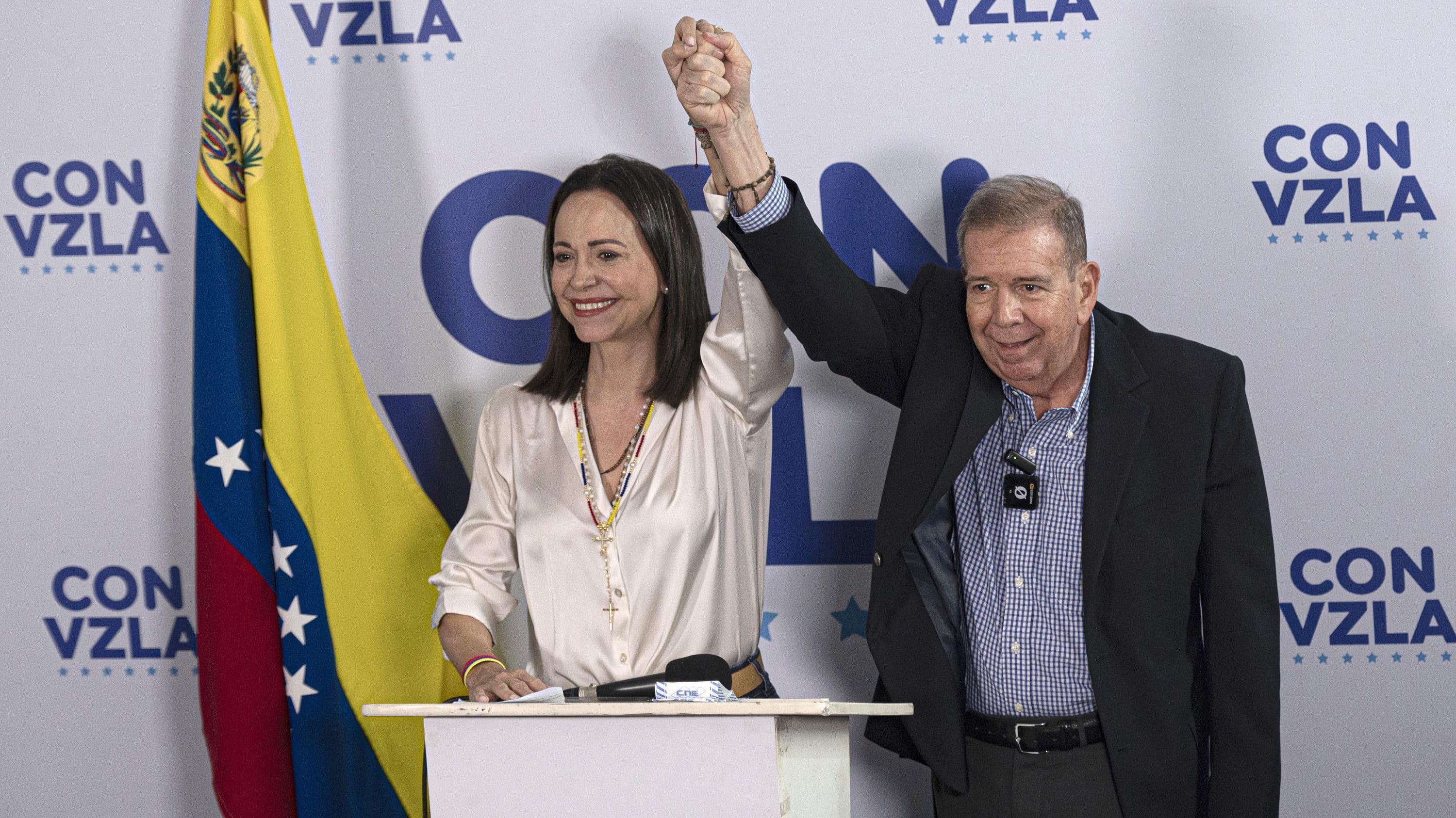 Maria Corina Machado y Edmundo González levantan sus brazos agarrados de la mano. En el fondo, una bandera de Venezuela y un logo que dice "Con Vzla"