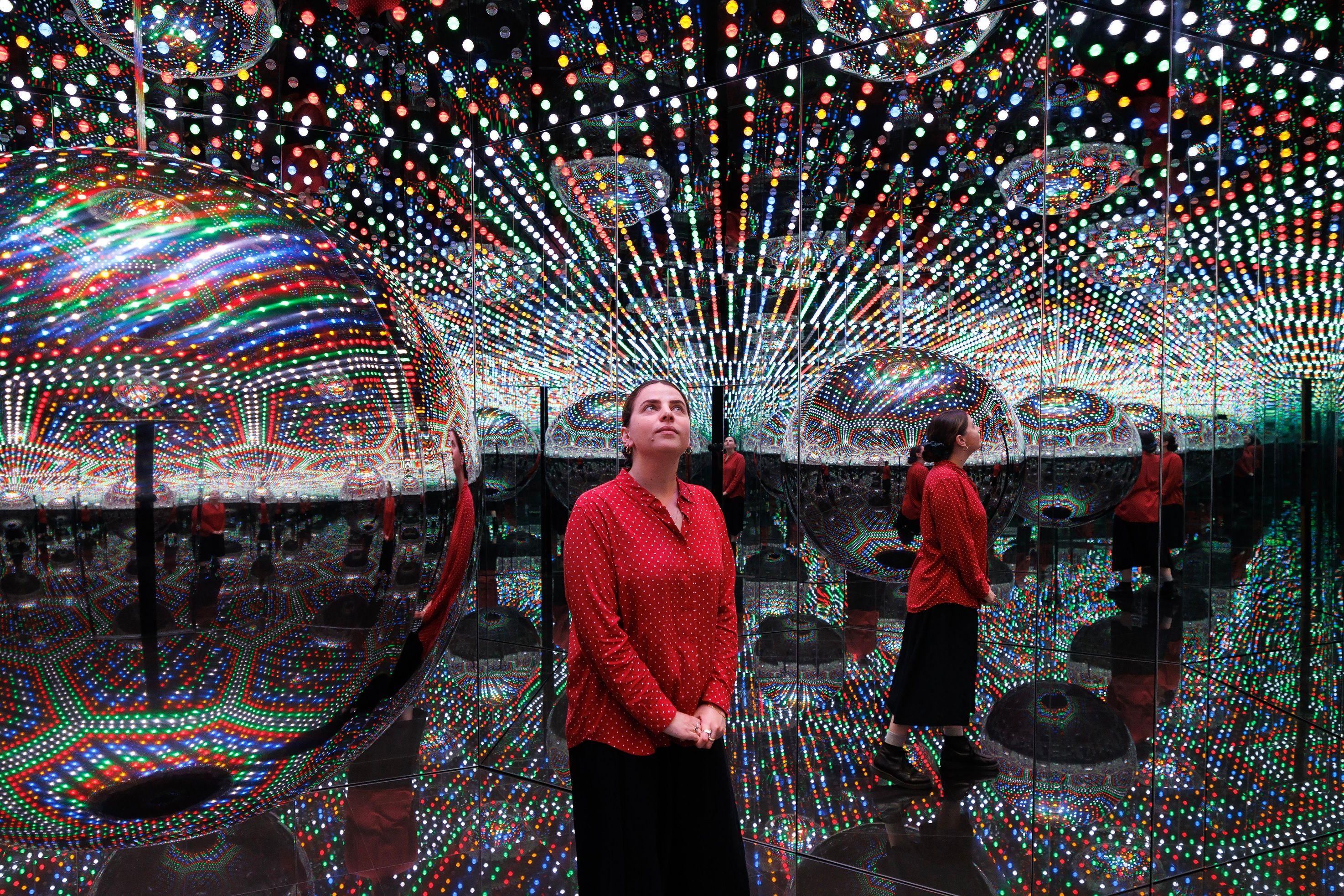 Una visitante de pie en una de las Salas Infinitas de Kusama, vista reflejada varias veces en espejos rodeados de luces.