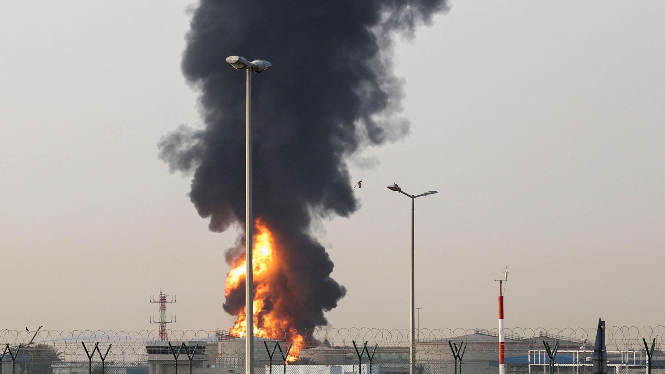 在阿拉伯聯合大公國杜拜，杜拜國際機場附近的一處區域升起滾滾濃煙與烈焰。畫面中可見數棟低矮建築，四周環繞著一道保全圍欄，似乎由盤繞的帶刺鐵絲或刀片刺網構成。