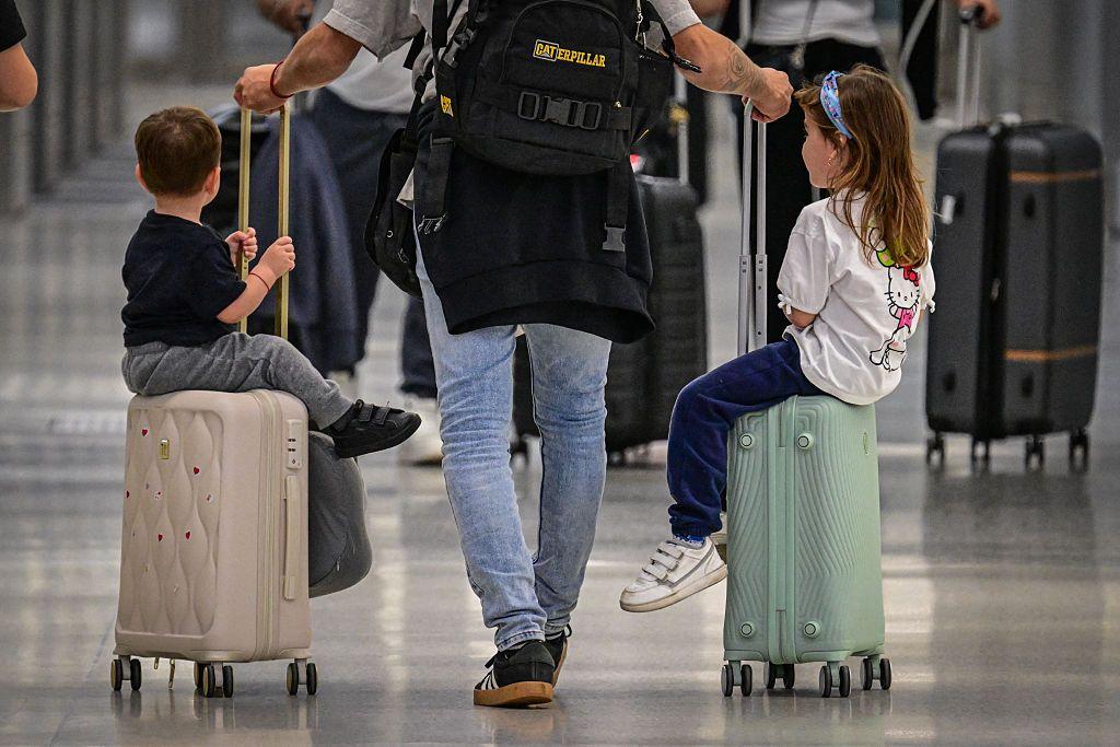 Un hombre lleva dos maletas de mano en las que están sentados dos niños.