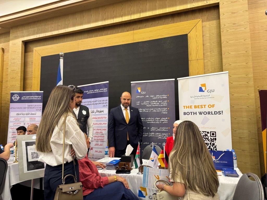 تُظهر الصورة مجموعة من الأشخاص يتفاعلون في ما يبدو أنه معرض أو فعالية. يتوسط الصورة رجل يرتدي بدلة رسمية وربطة عنق وهو الدكتور مالك الشرايري، يقف خلف طاولة عرض تحتوي على مواد ترويجية وشعارات