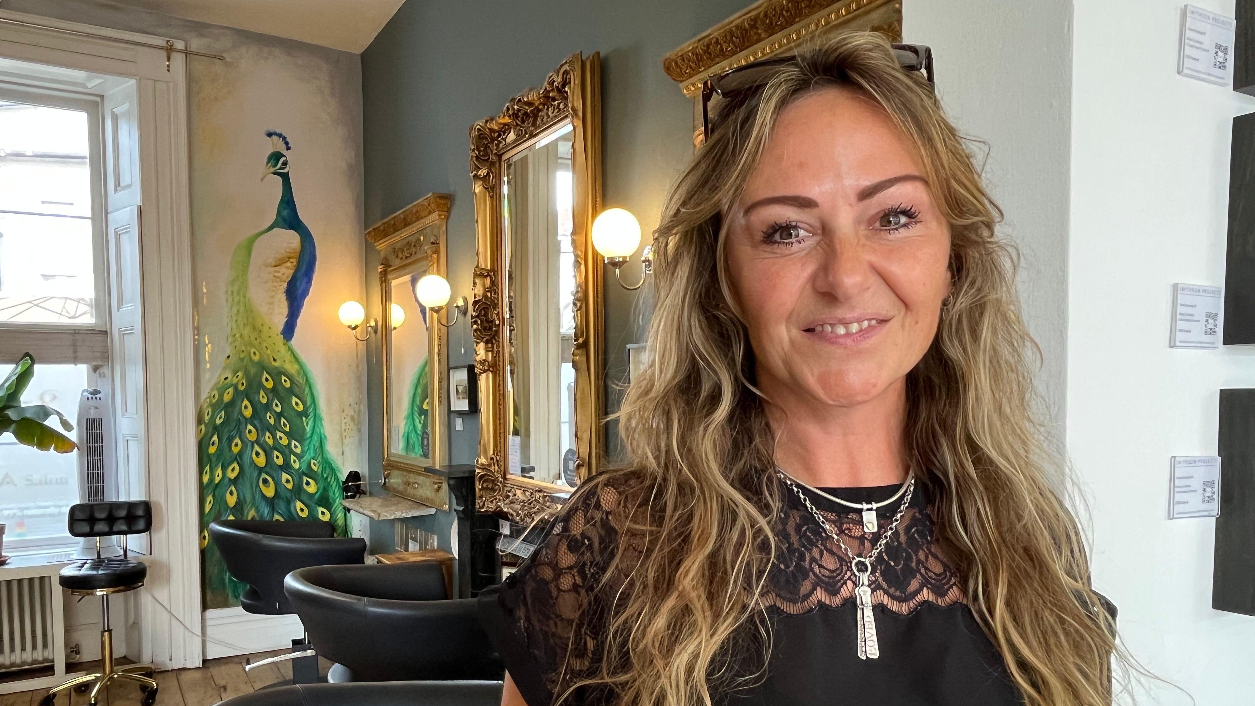 En el interior de una peluquería, una mujer de cabello rubio y ondulado sonríe directamente a la cámara, con los ojos brillantes. Al fondo, un cuadro de un pavo real adorna una pared, junto a grandes espejos con marcos dorados y sillones negros para los clientes.