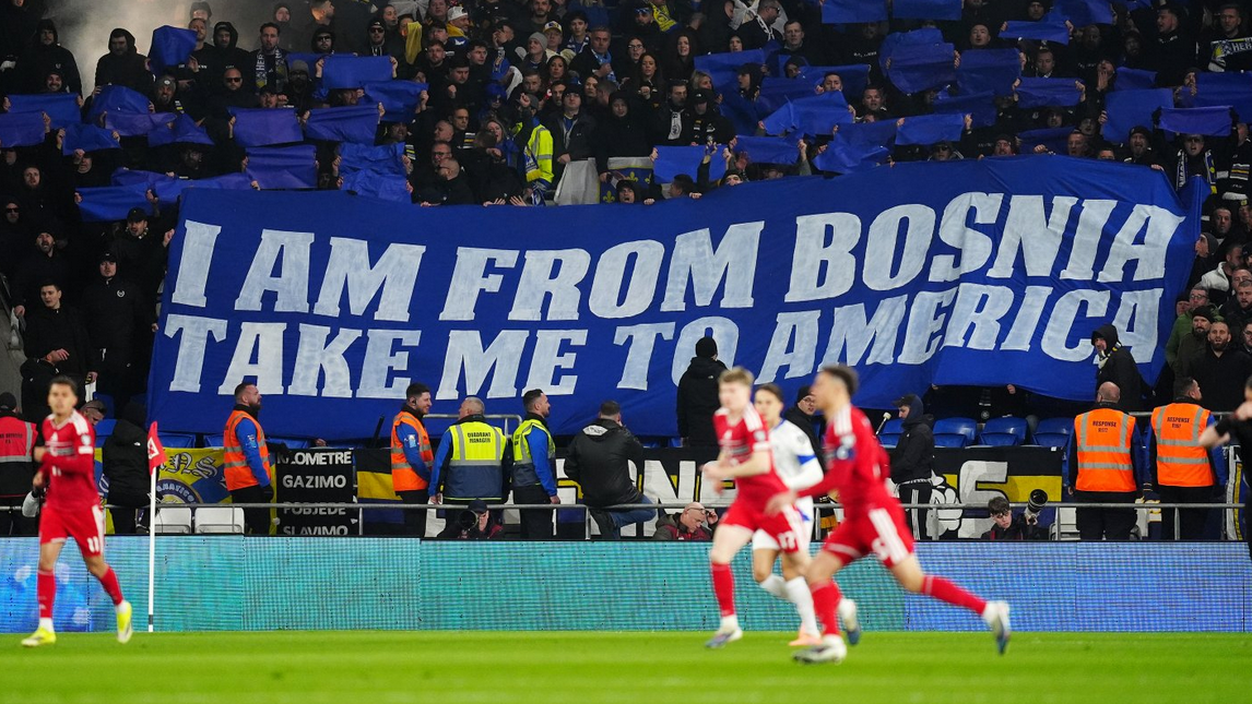 Navijači BiH drže ogroman transparent na plavom platnu na kojem belim slovima piše na engleskom jeziku stih pesme Dubioza Kolektiva - I am from Bosnia, take me to America, ispred 3 igrača u crvenom dresu trče na terenu