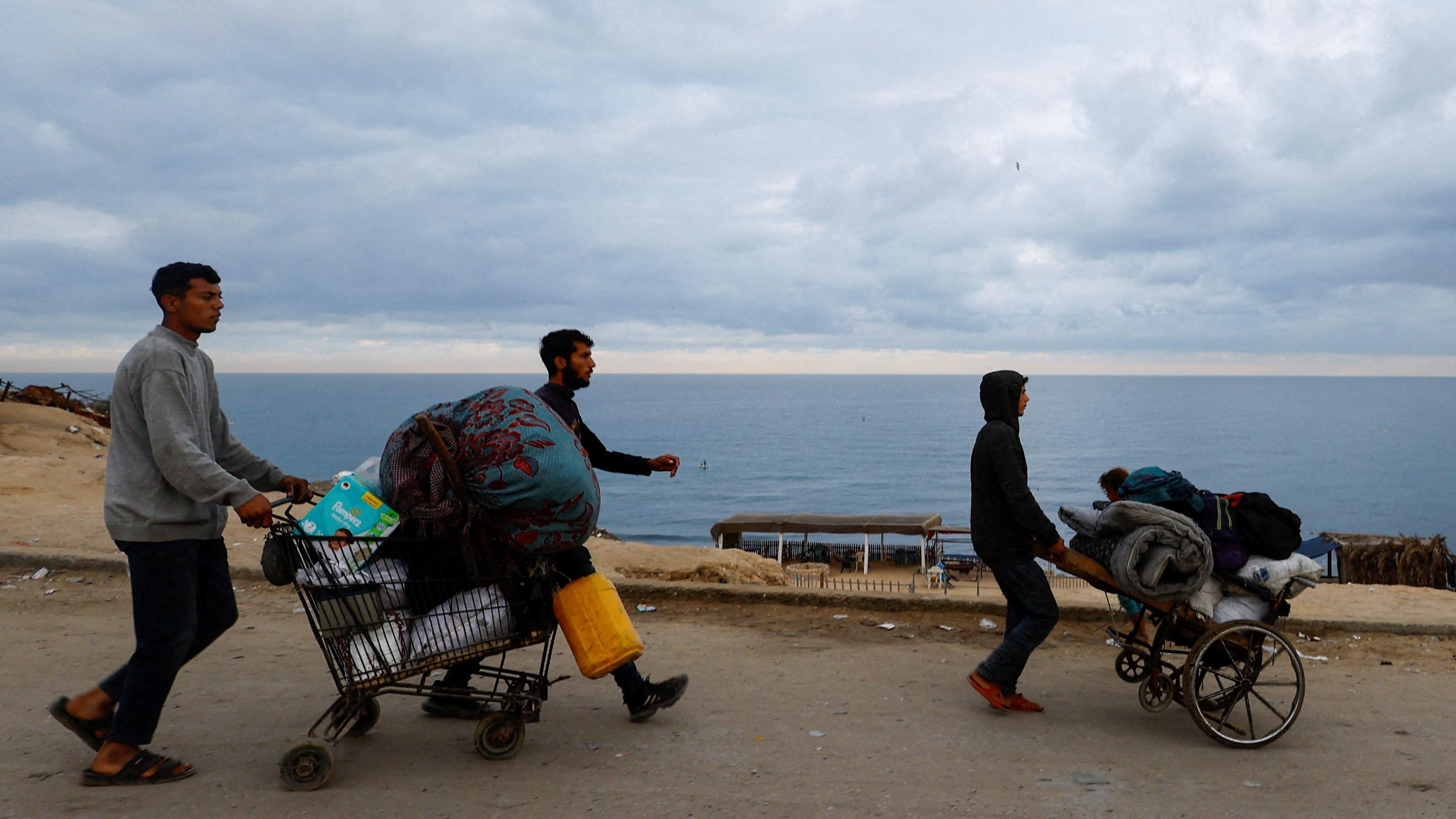 Tres hombres empujan un carrito de la compra cargado con enseres personales. Al fondo se ve el mar.