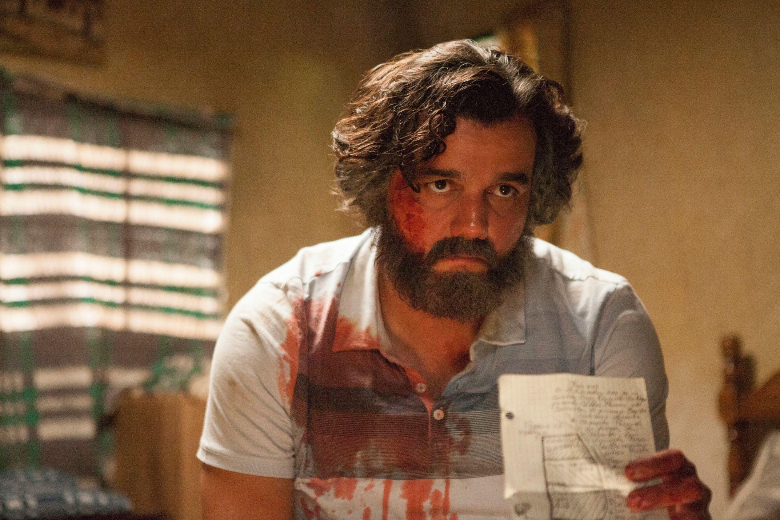 O ator Wagner Moura aparece como Pablo Escobar, com o rosto e a camisa manchados de sangue, segurando um papel com anota&ccedil;&otilde;es enquanto encara a c&acirc;mera com express&atilde;o tensa em cena da s&eacute;rie Narcos.