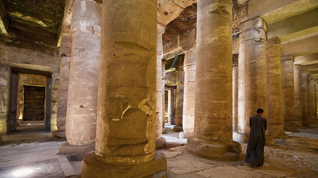 عواميد ضخمة ورجل بعباءة داكنة اللون، يمشي في ممر جانب العواميد الضخمة في معبد أبيدوس في سوهاج في مصر