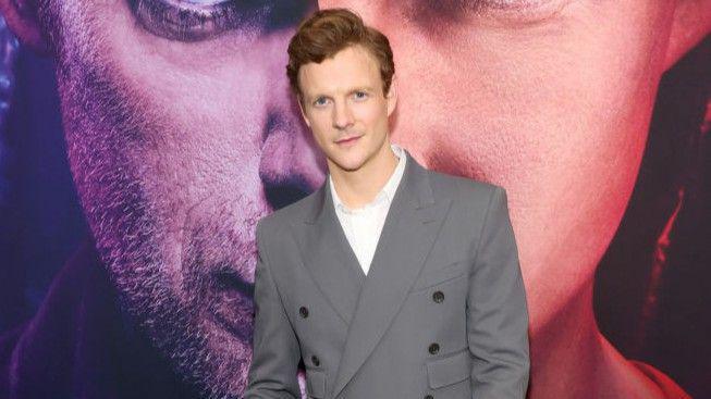 Patrick Gibson em um terno cinza em um tapete vermelho.