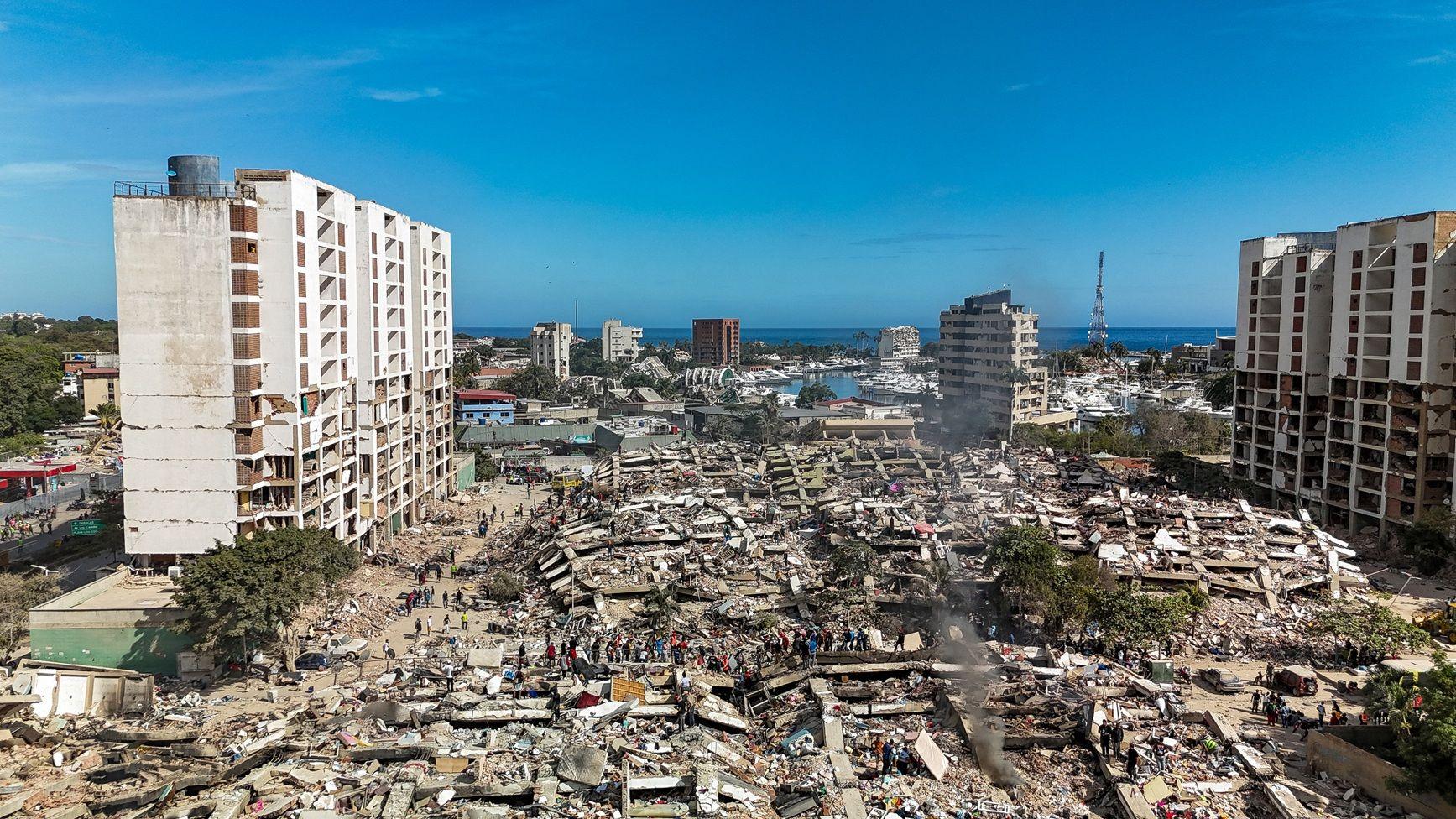Dos edificios, gravemente afectados, están de pie. En el centro, se ven edificaciones totalmente colapsadas. Grupos de personas caminan en el área. El cielo azul está totalmente despejado. 