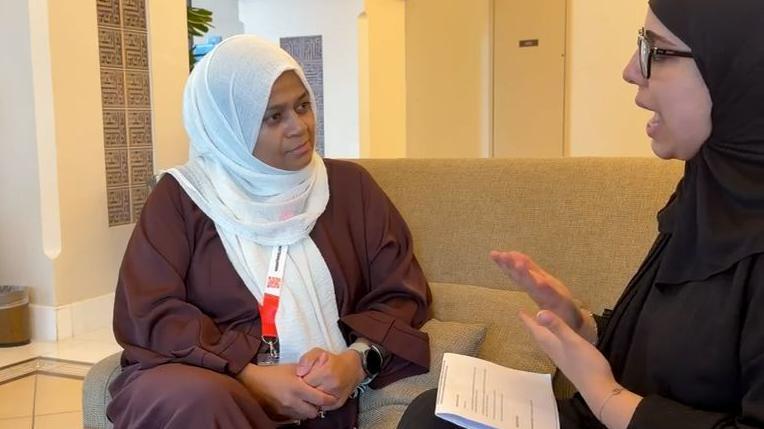 امرأتان ترتديان زياً إسلامياً تجلسان على أريكة متقابلتين. تظهر زينام على اليمين مرتدية حجاباً وعباءة باللون الأسود، وتستخدم لغة الإشارة للتواصل مع جاني، التي تجلس على اليسار مرتدية عباءة بنية وحجاباً أبيض، وتضع حول عنقها شارة تعريف باللونين الأحمر والأبيض.