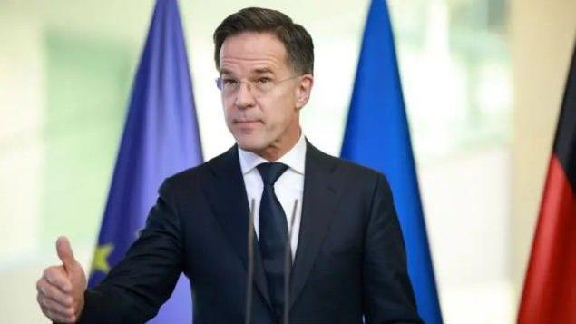  الأمين العام للناتو مارك روته مرتدياً بدلة رسمية ويرفع إحدى يديه.