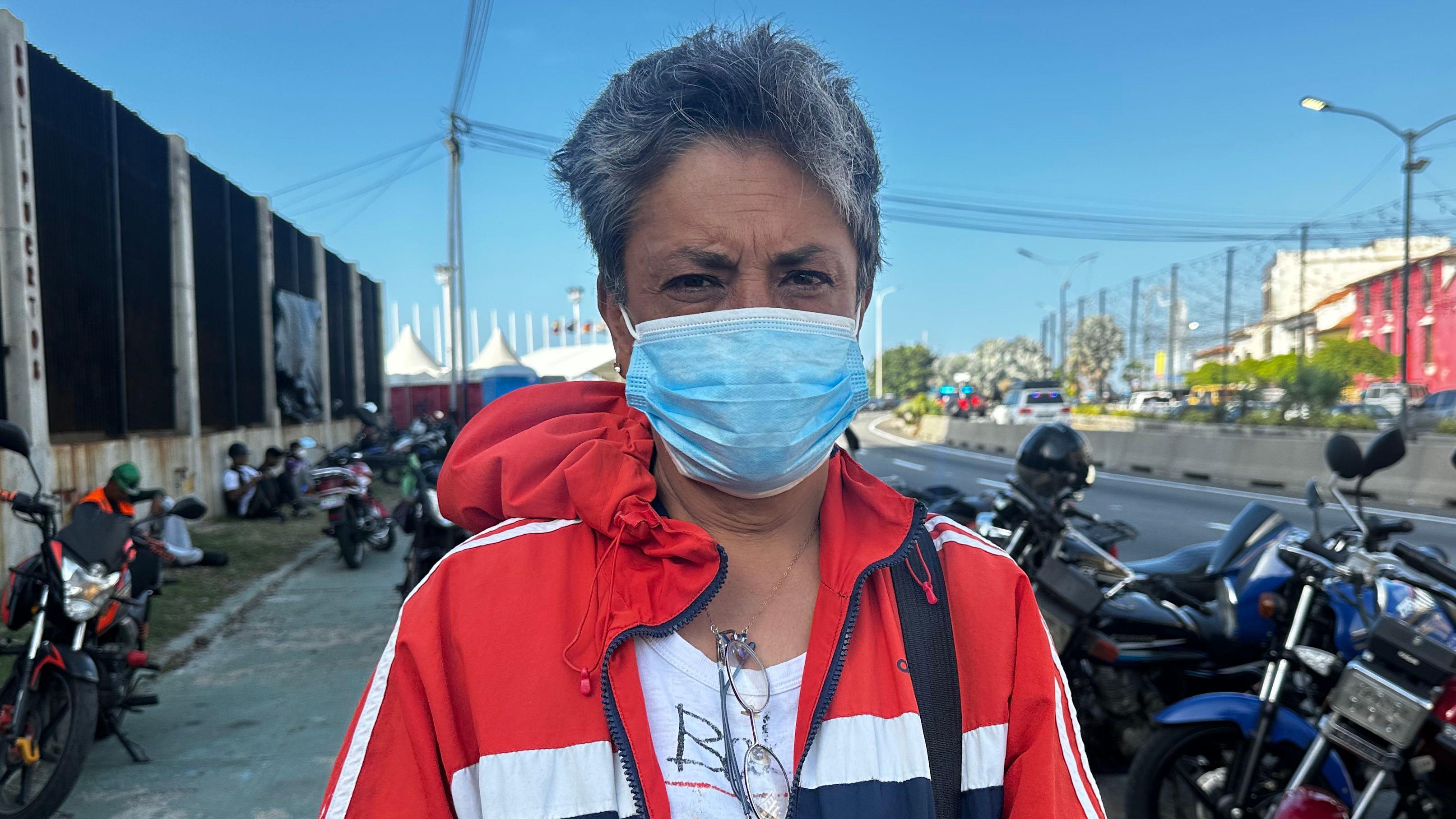 Modesta Alemán con un tapabocas azul frente a Los Silos. Lleva una chaqueta roja con rayas azules y blancas. 
