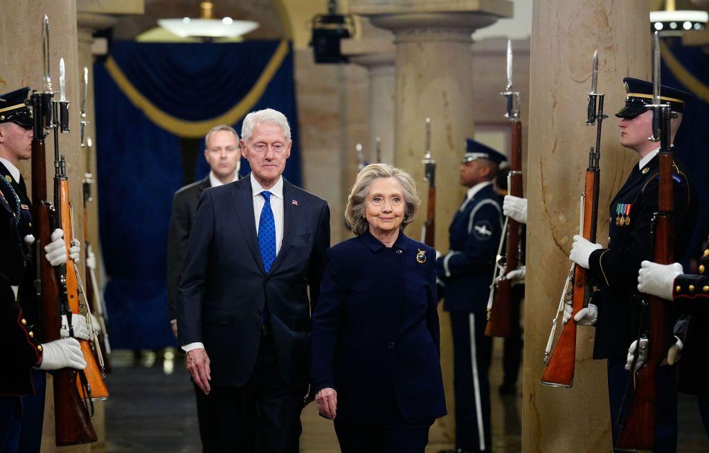 Bill e Hillary caminhando um lado do outro. Ao lado h&aacute; v&aacute;rios guardas