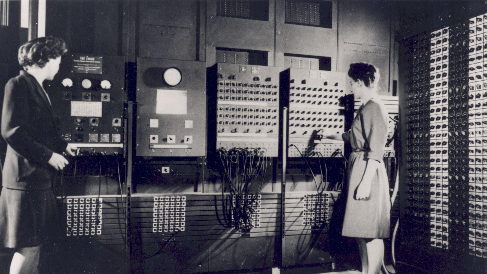Fotografia em preto e branco mostra duas mulheres diante de um grande computador de primeira gera&ccedil;&atilde;o, cercado por pain&eacute;is de controle, cabos e fileiras de componentes eletr&ocirc;nicos, em uma sala de processamento de dados.