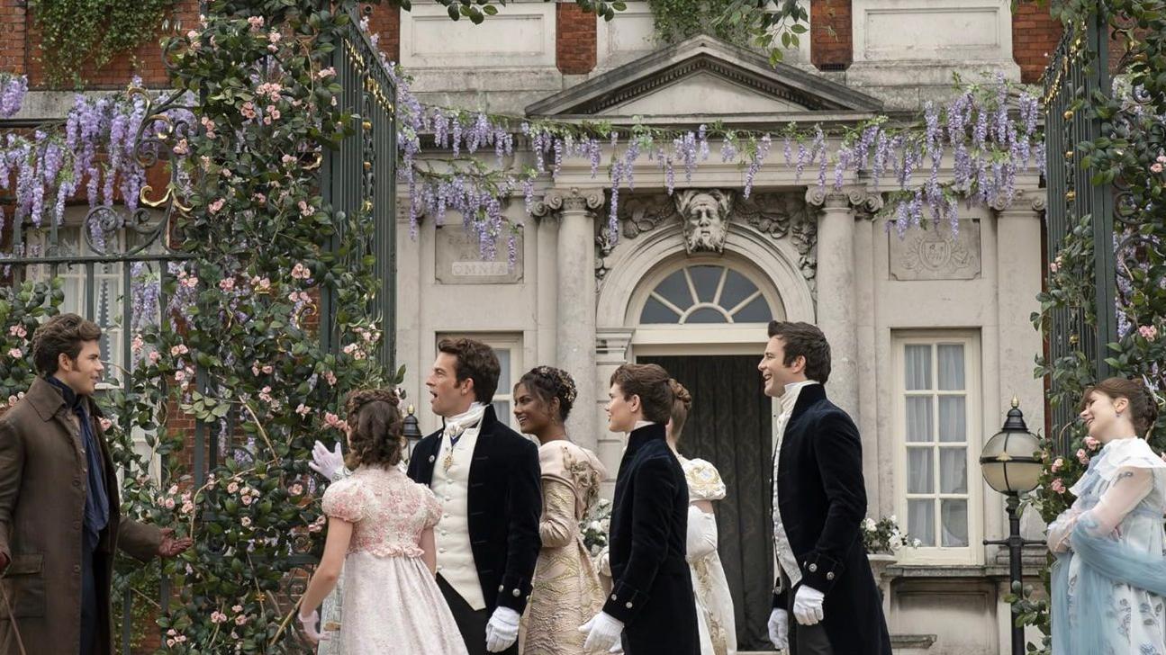La familia Bridgerton en la puerta de su cara, la caracterísitca Ranger's House, de estilo georgiano, con las flores violetas colgantes adornando las paredes. 