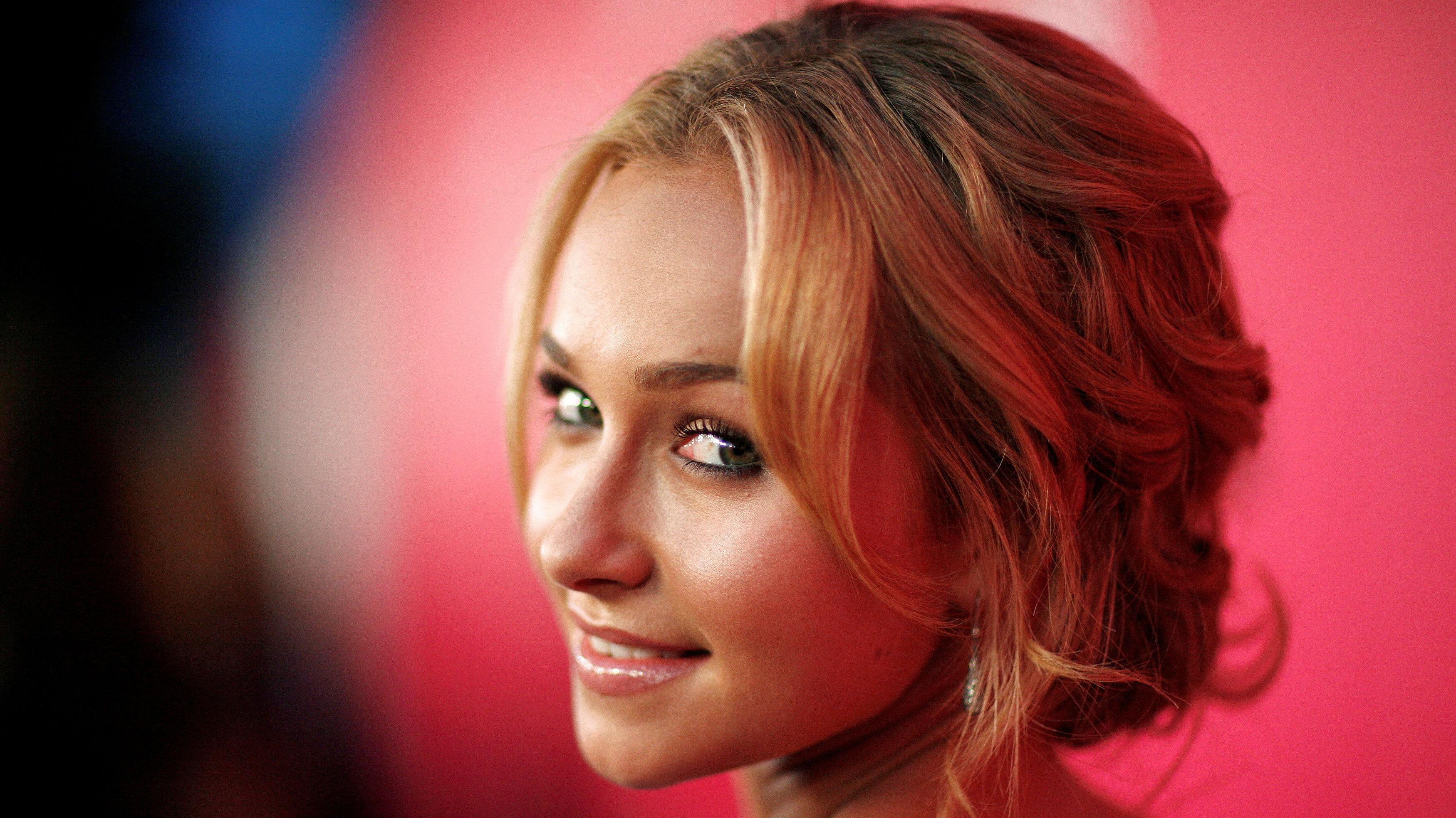 La cabeza de Hayden Panettiere se ve girada para mirar hacia atrás y sonreír suavemente, frente a un fondo rojo.