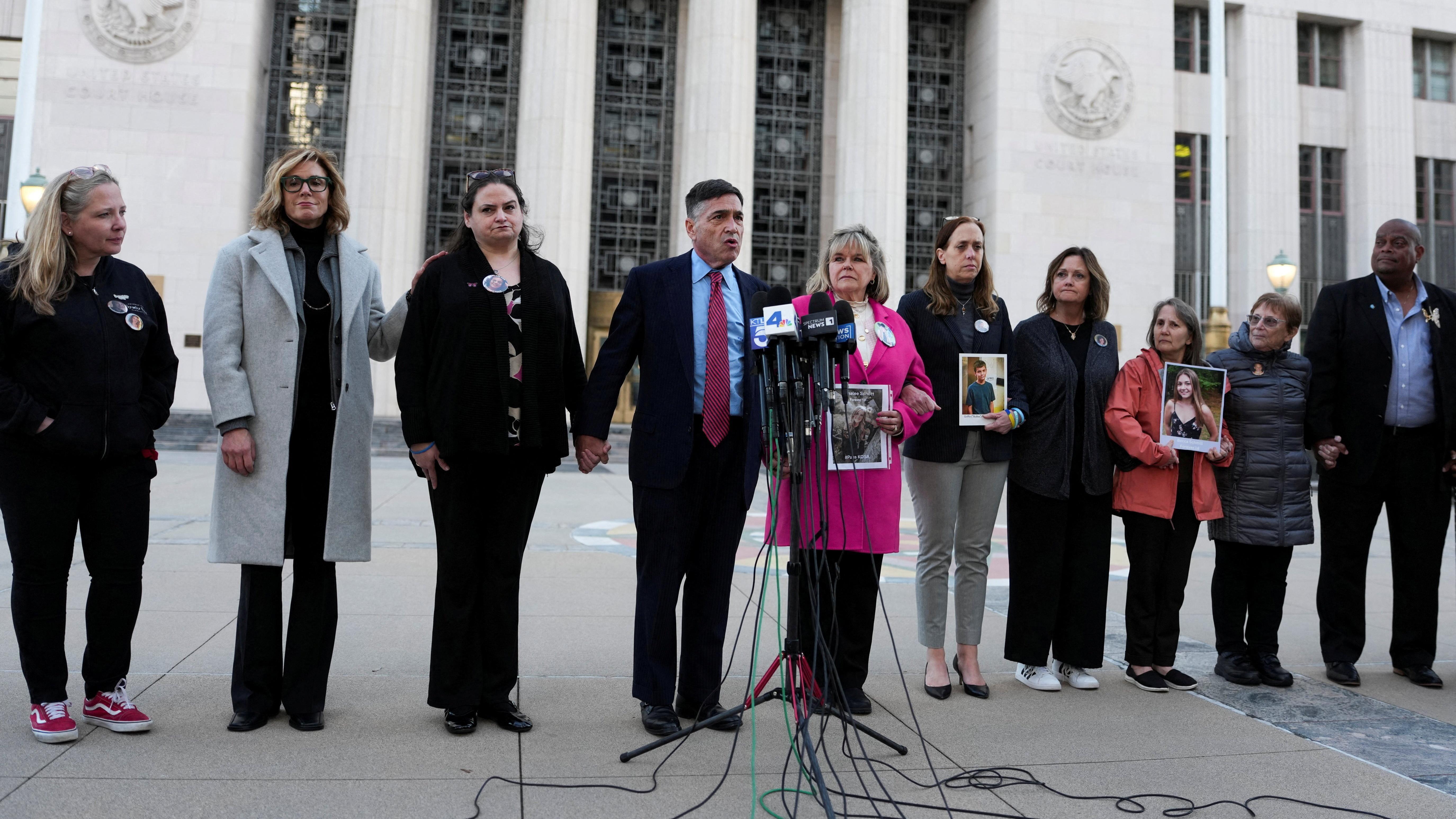 صورة تظهر عدداً من الأشخاص يقفون مصطفين، بعضهم يحمل صوراً لأطفال، ويتقدمهم رجل يتحدث أمام مايكروفون.