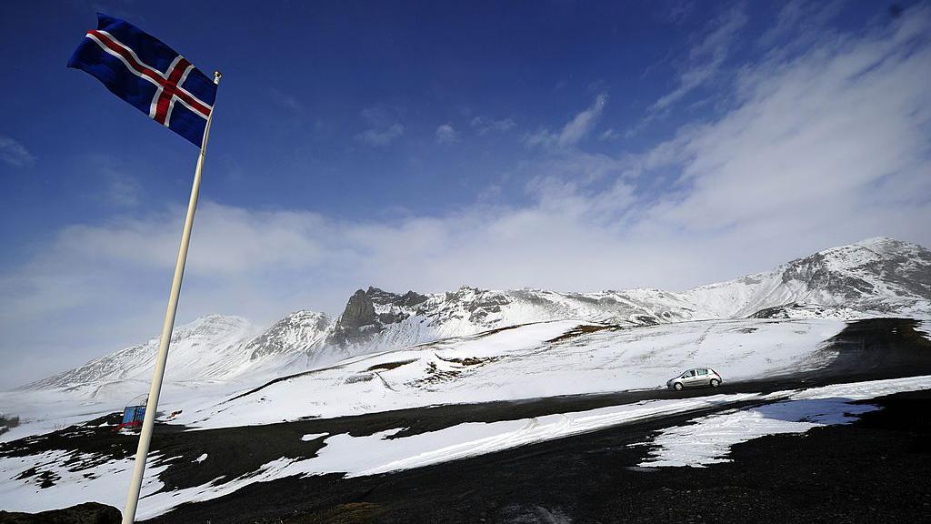 Uma bandeira islandesa tremula no alto de um mastro na encosta de uma montanha nevada.