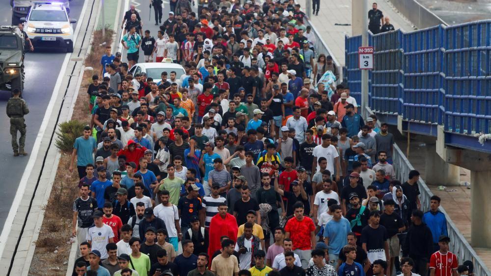 Um grande grupo de migrantes, principalmente homens, sendo escoltado pela polícia ao longo de uma passarela. 