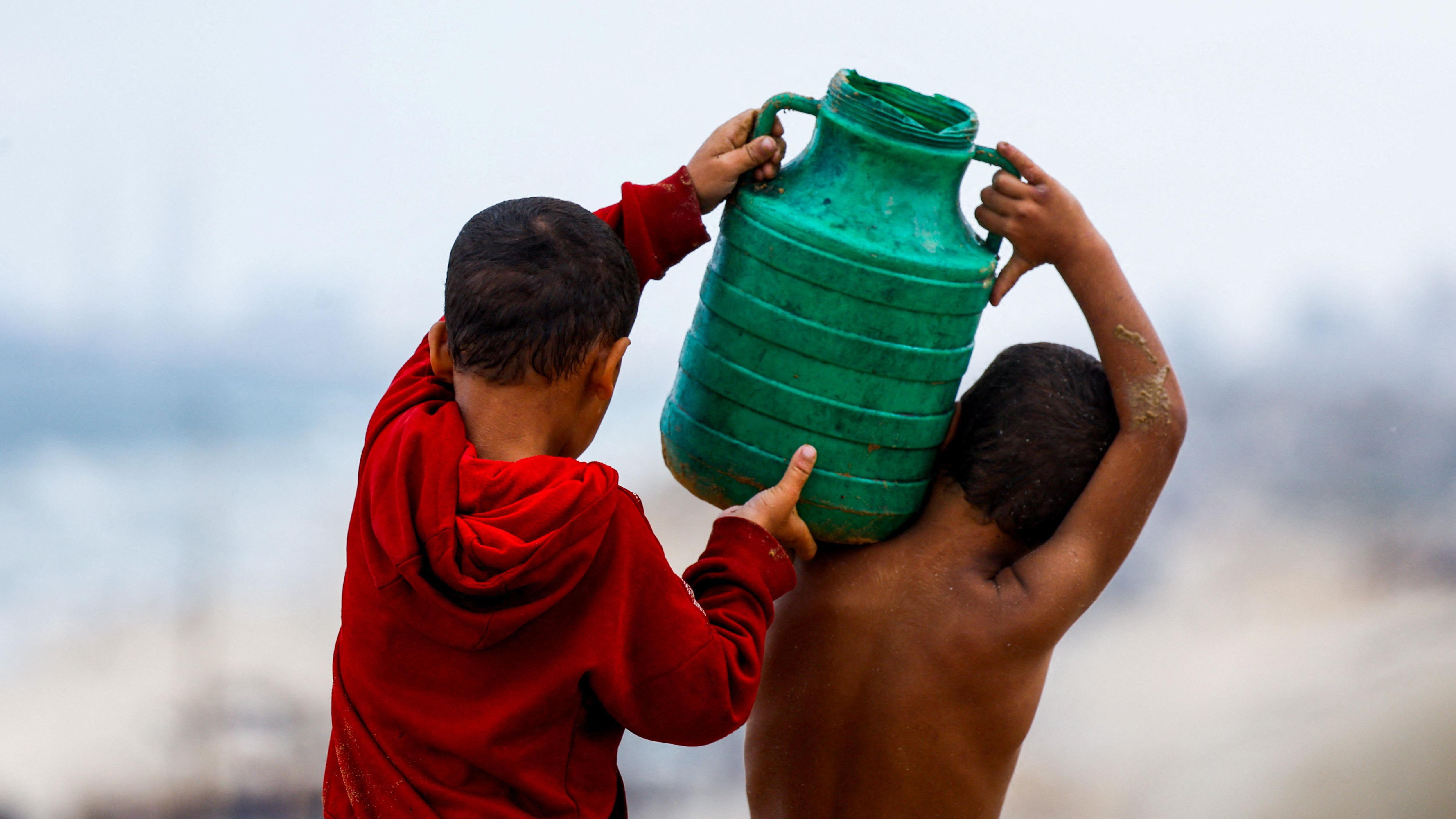 Dos niños llevan al hombro una garrafa de plástico verde de agua.