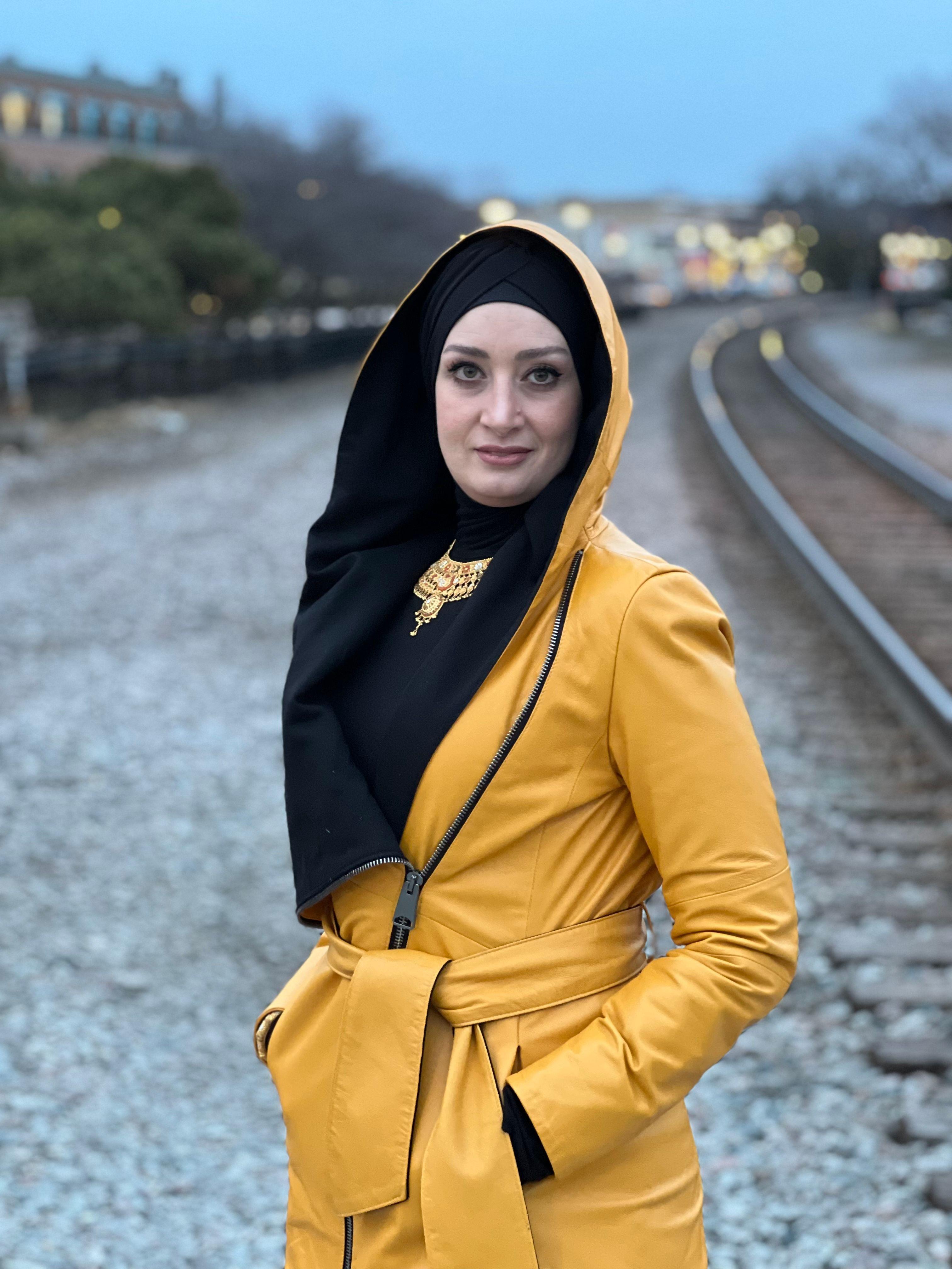 سيدة ترتدي حجاباً أسود ومعطفاً أصفر تضع يديها في جيبي المعطف وتقف على ما يبدو بالقرب من خطوط السكة الحديدية