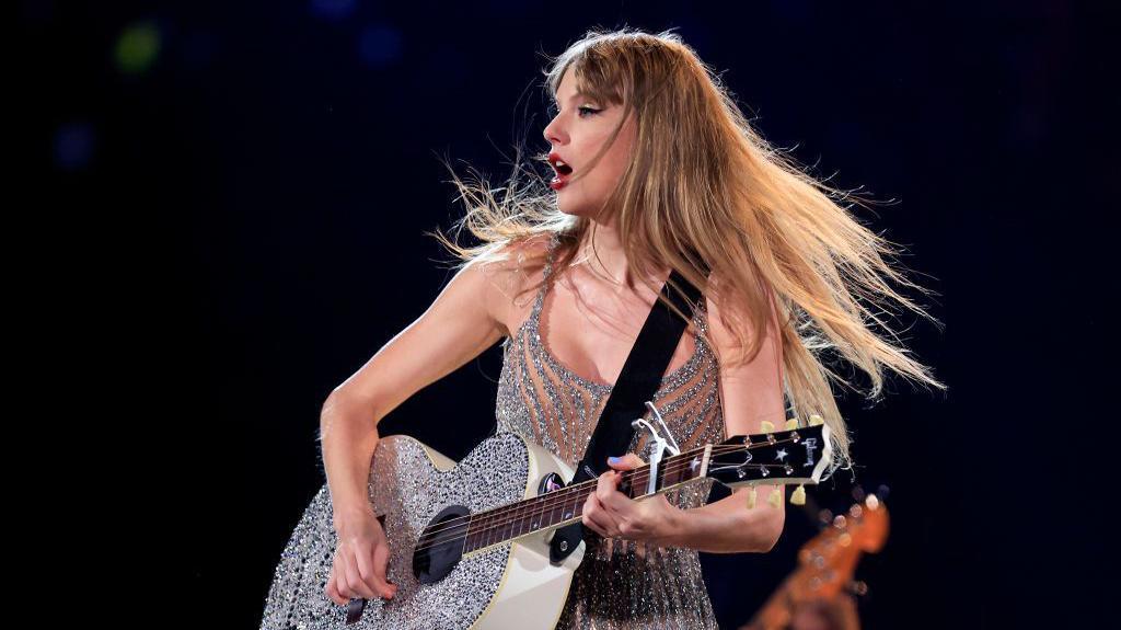 Taylor Swift em apresenta&ccedil;&atilde;o ao vivo, tocando um viol&atilde;o prateado coberto de brilho enquanto canta. Com os cabelos esvoa&ccedil;ando durante o movimento e vestindo um figurino prateado com franjas, ela aparece iluminada pelos refletores do palco.