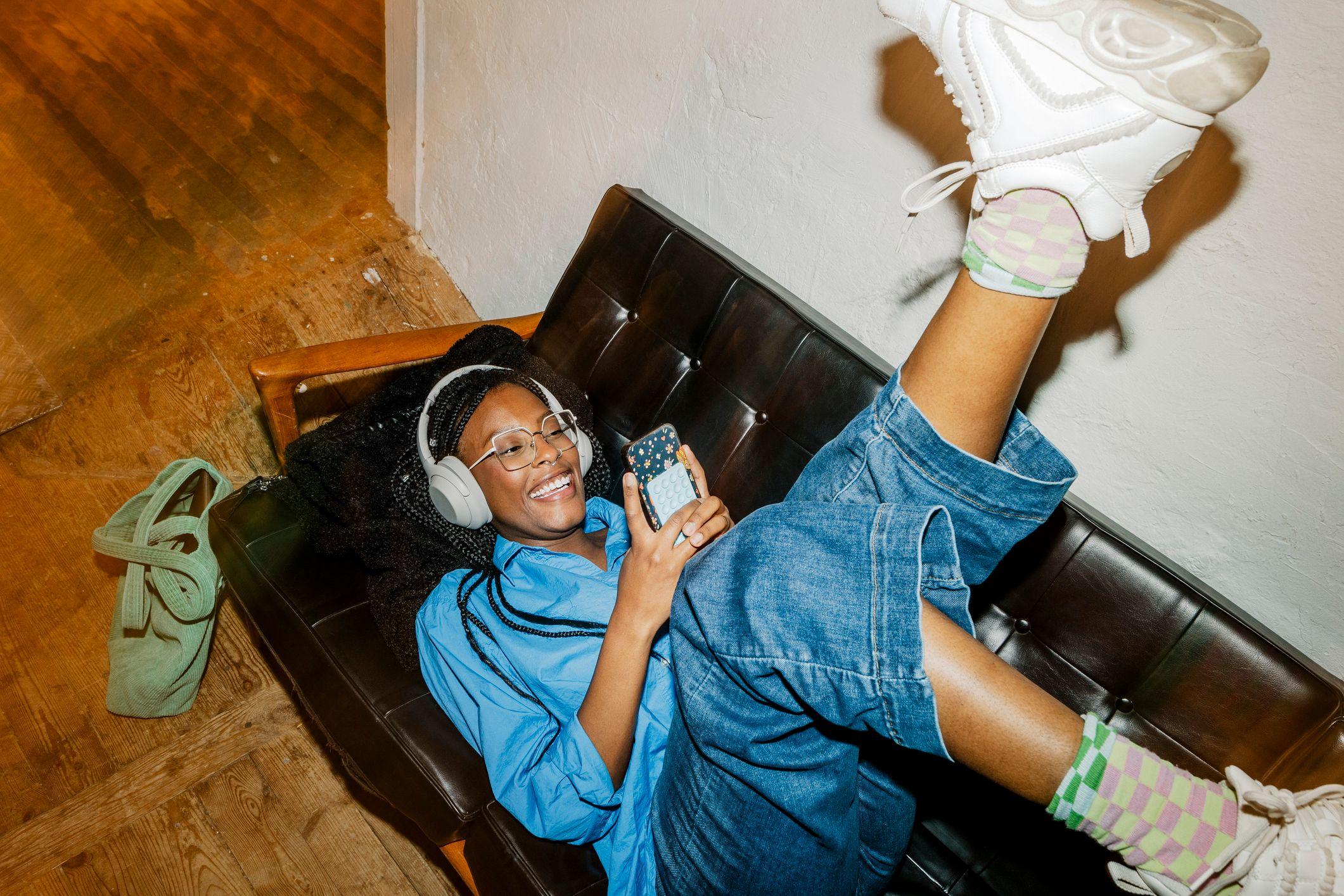 Mulher com fone de ouvidos deitada em um sof&aacute; com as pernas para cima, claramente se divertindo enquanto olha seu telefone celular.