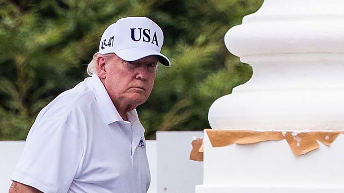 Donald Trump, vestido con polo blanco, pantalón negro y una gorra blanca con el lema "USA", junto a una gran columna blanca. 