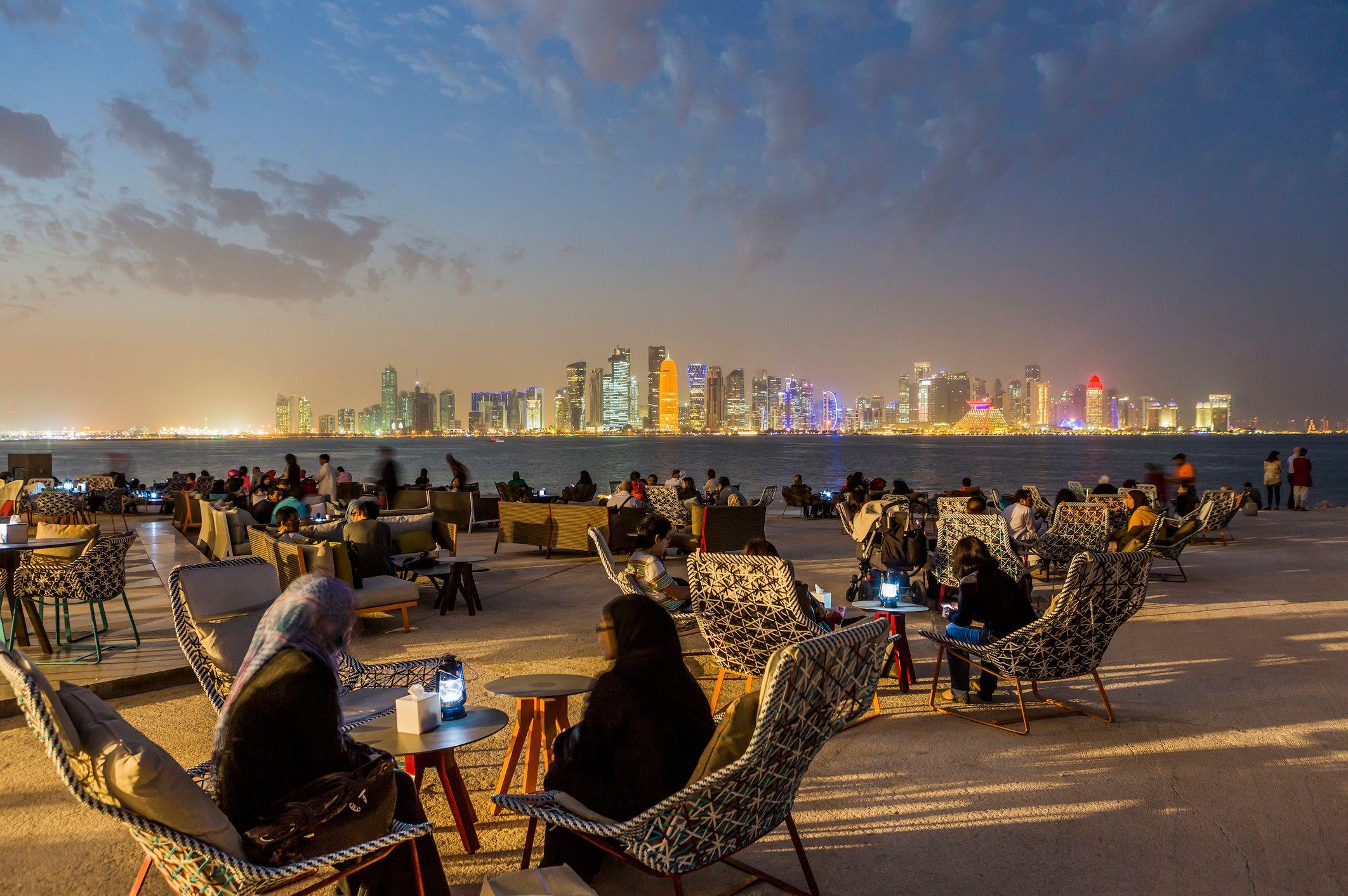 Pessoas sentadas em mesas de restaurantes, com prédios iluminados atrás, ao anoitecer
