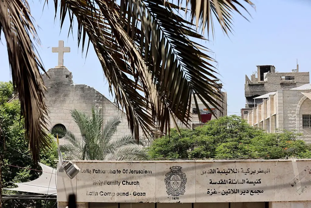 صليب على كنيسة متضررة وأمامها مكتوب على لوحة أسمنتية "البطريركية اللاتينية كنيسة السائلة المقدسة مجمع دير اللاتين -غزة"، باللغتين العربية والإنجليزية. وهناك عدد من الأشجار. الصورة التُقطت نهاراً.