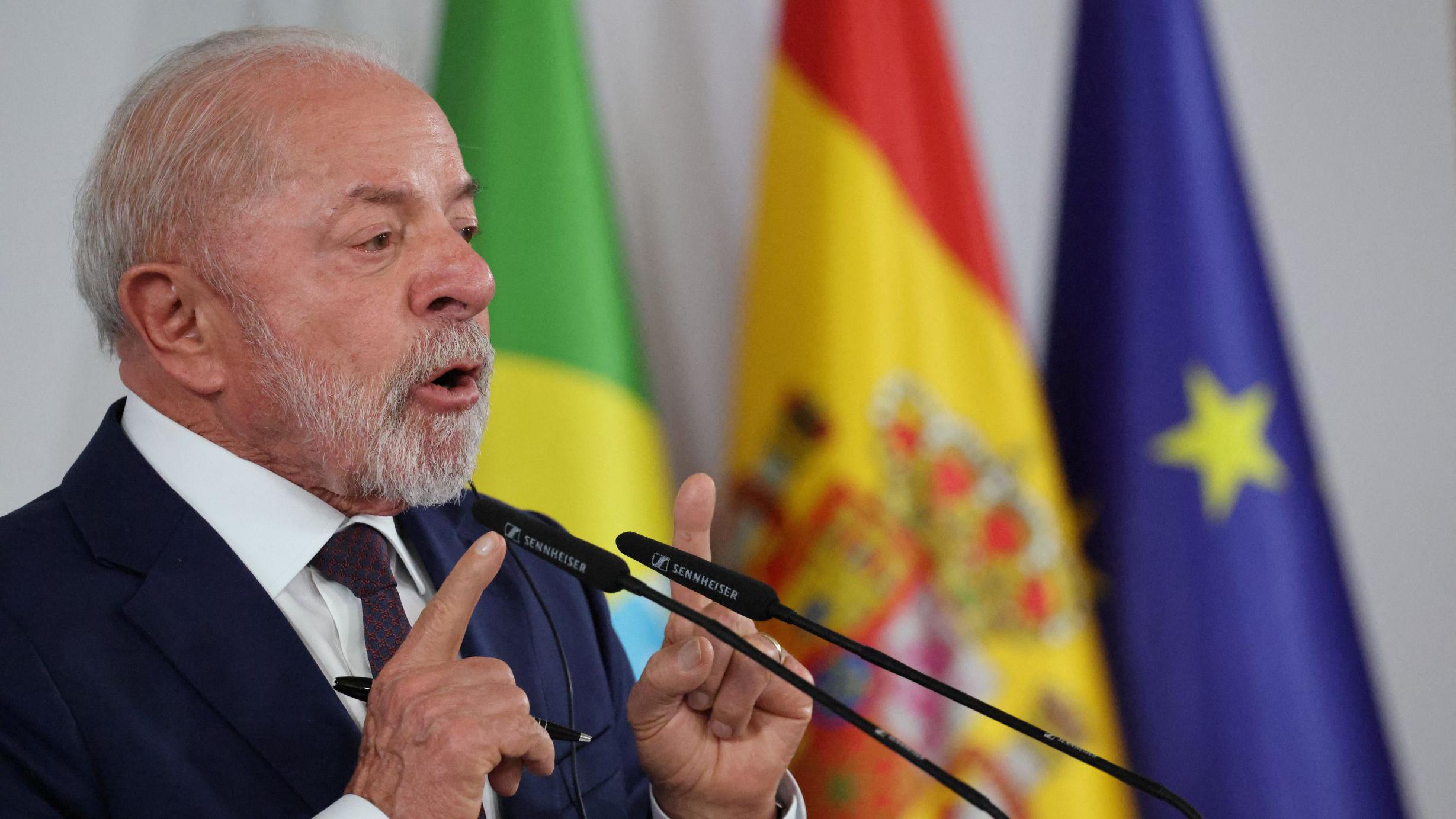 O presidente Lula, de barba e cabelo grisalhos, veste terno escuro, camisa branca e gravata, enquanto discursa em um p&uacute;lpito com dois microfones &agrave; frente. Ele gesticula com as m&atilde;os levantadas, apontando os dedos indicadores. Ao fundo, aparecem bandeiras desfocadas &mdash; do Brasil, da Espanha e da Uni&atilde;o Europeia.