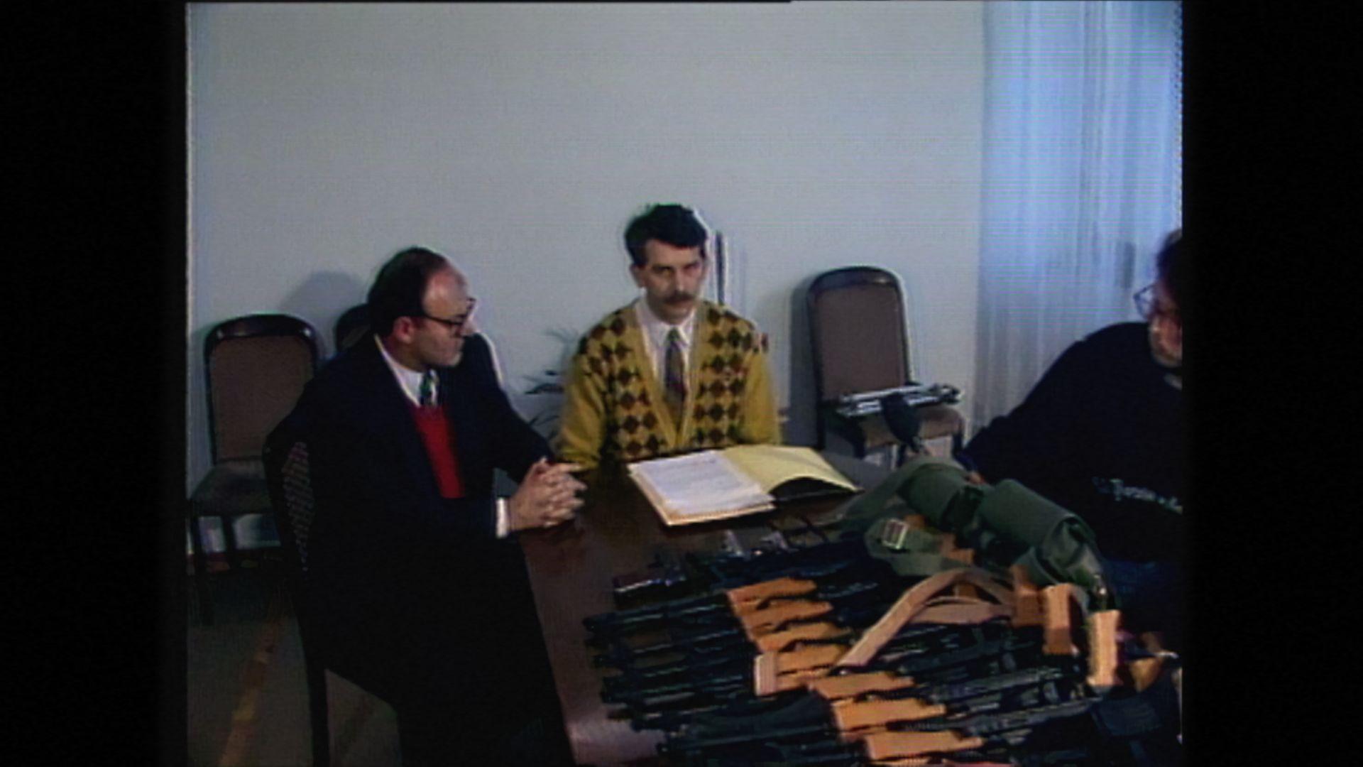 Dva muškarca sede ispred velike količine oružja, jedan ima brkove