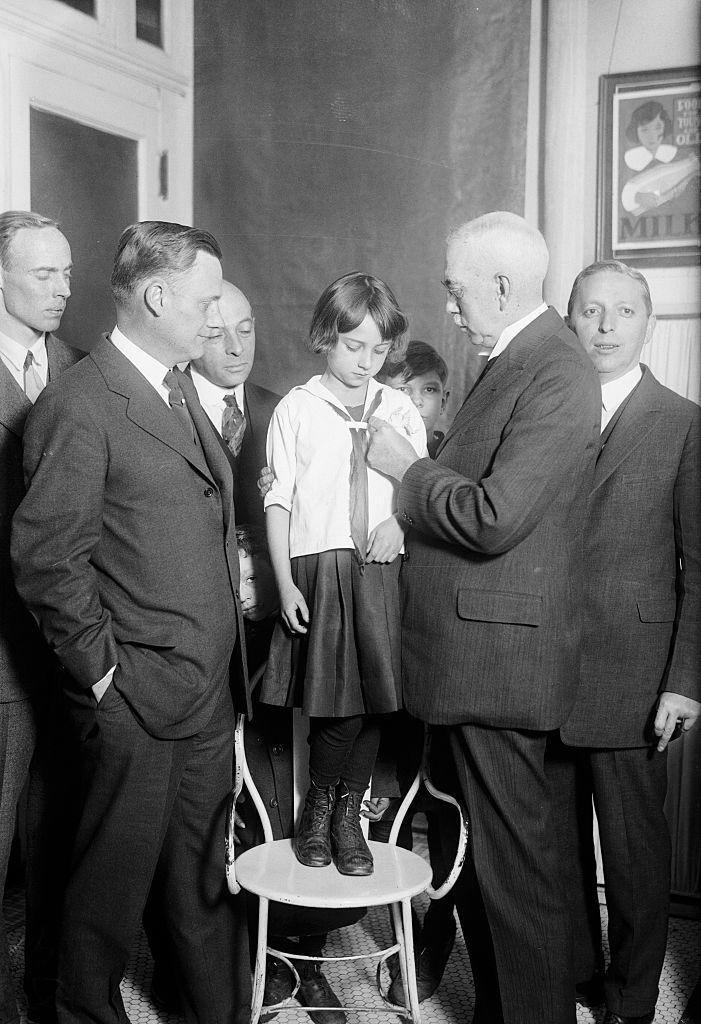 ي نيويورك، بتاريخ 28 أكتوبر عام 1925، تظهر فتاة وهي تتلقى التطعيم ضد الدفتيريا. في الصورة، يقوم الدكتور فرانك ج. موناغان، مفوض الصحة بمدينة نيويورك، بوضع شارة التطعيم على قميص الفتاة روز غورملي، التي كانت قد تلقت تطعيمها ضد الجدري وحصّنت ضد الدفتيريا بحقنة من مضاد السم.