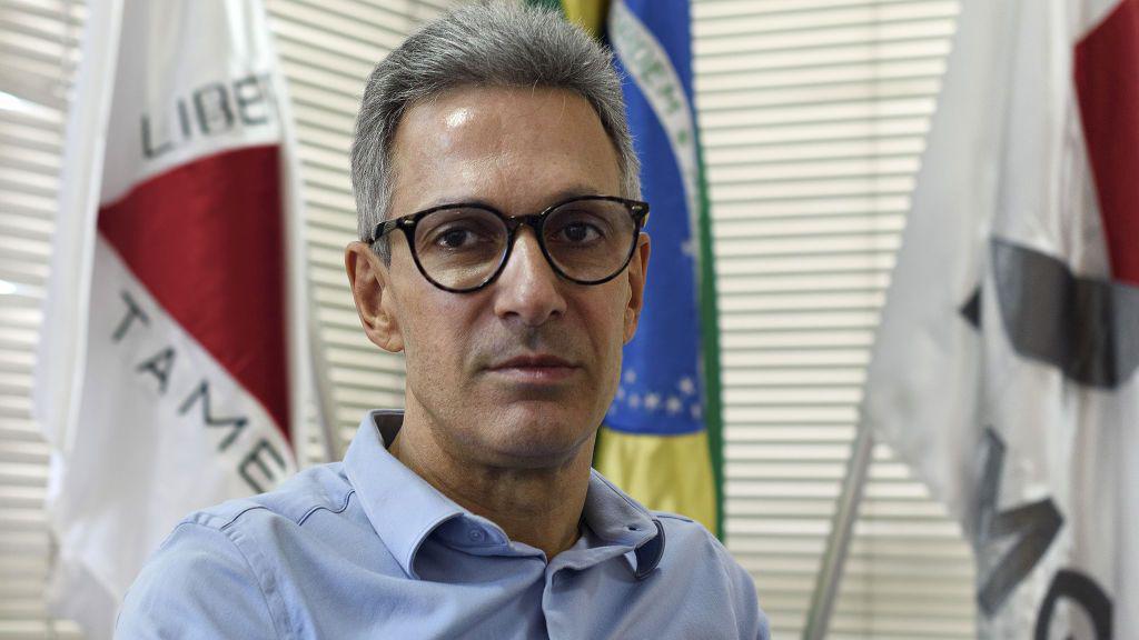 Romeu Zema &eacute; um homem branco e usa &oacute;culos de grau com arma&ccedil;&atilde;o preta. Ele est&aacute; com uma camisa azul clara que tem uma bandeira de Minas Gerais. 