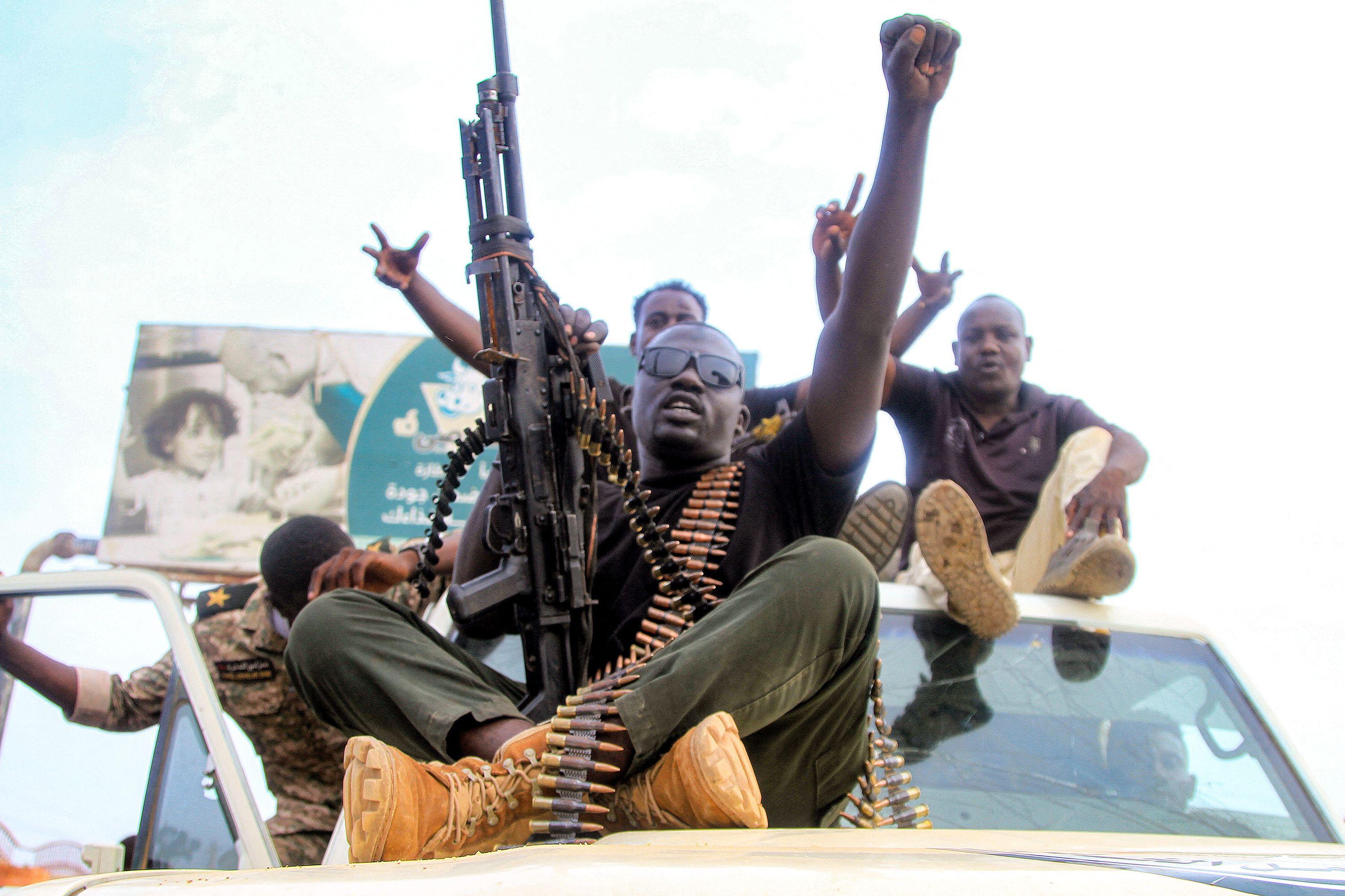 Varios hombres, el primero armado con una ametralladora y con una cinta de balas sentados en el capó de una camioneta. 