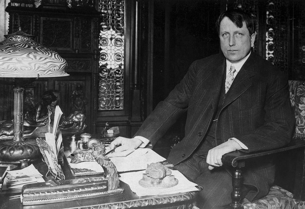 William Randolph Hearst, sentado en un gran escritorio de madera con varios objetos dorados encima.