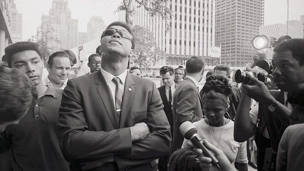 محمد علي يرتدي نظارة سوداء وينظر إلى السماء محاطاً بالصحافيين