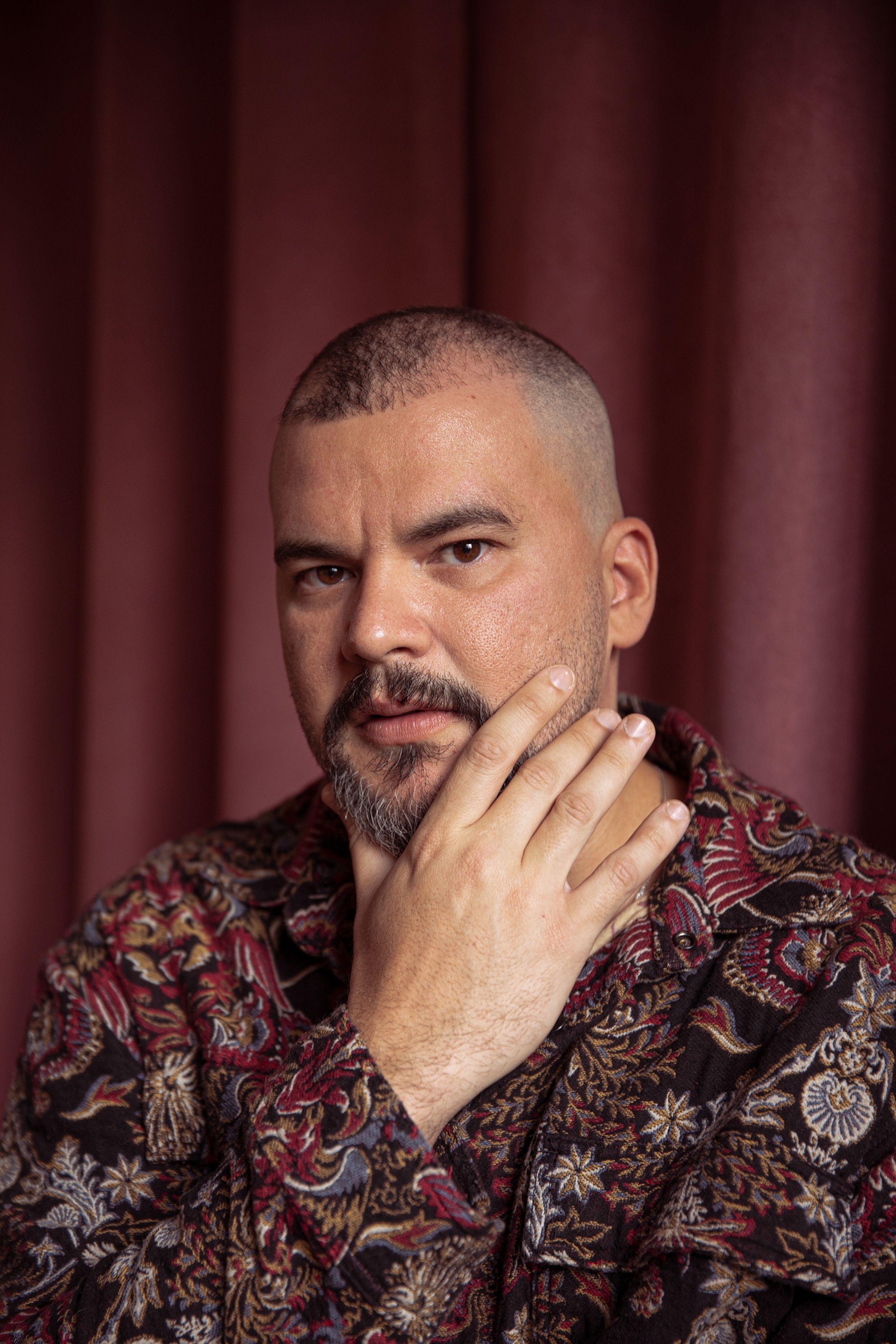 Retrato de um homem de cabe&ccedil;a raspada e barba curta, com express&atilde;o s&eacute;ria, olhando para a c&acirc;mera. Ele apoia a m&atilde;o no rosto, tocando a bochecha, e veste uma camisa estampada em tons de vermelho, dourado e preto. Ao fundo, h&aacute; uma cortina vermelha que cria um contraste quente com a ilumina&ccedil;&atilde;o suave do retrato.