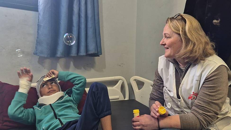 Katrin Glatz Brubakk junto a un niño herido en una cama de hospital soplando pompas de jabón
