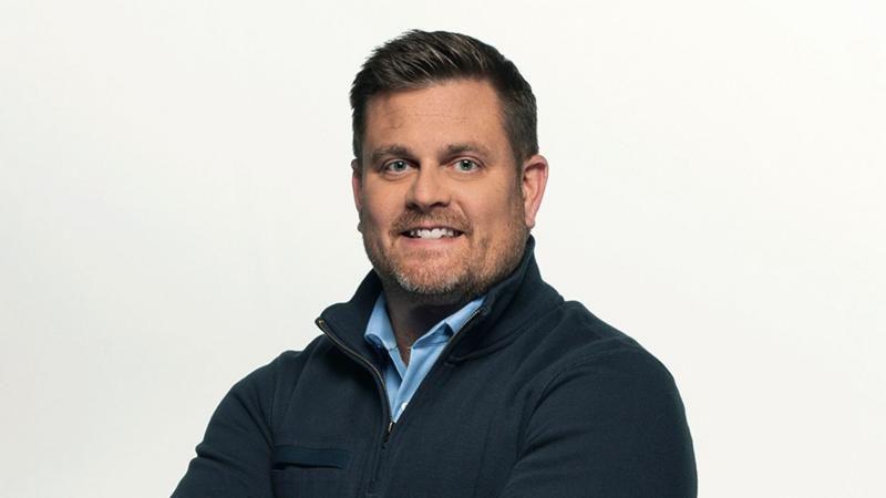 A foto mostra um homem, Brian Thompson, sorrindo, de bra&ccedil;os cruzados, usando uma camisa azul-clara sob uma jaqueta escura, diante de um fundo branco.