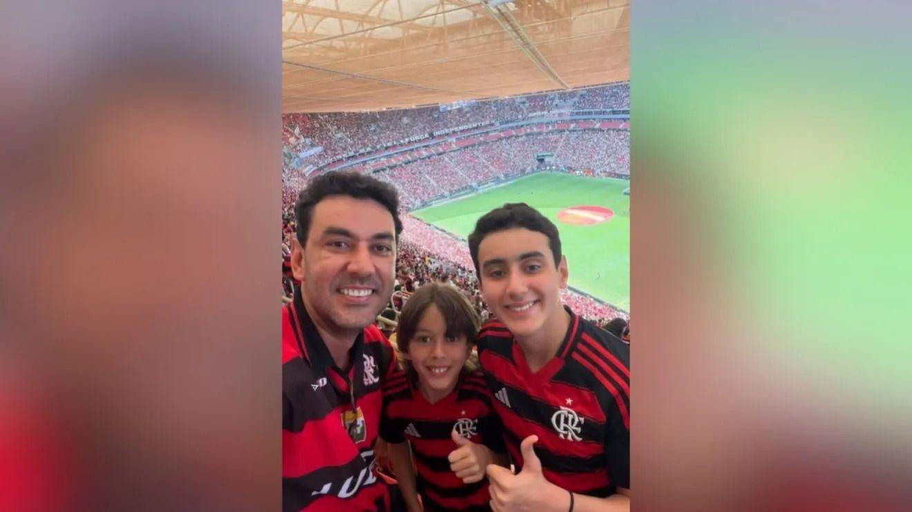 Thales Machado com os dois filhos, todos vestem camisas do Flamengo e sorriem olhando para a c&acirc;mera, na torcida de um est&aacute;dio de futebol lotado