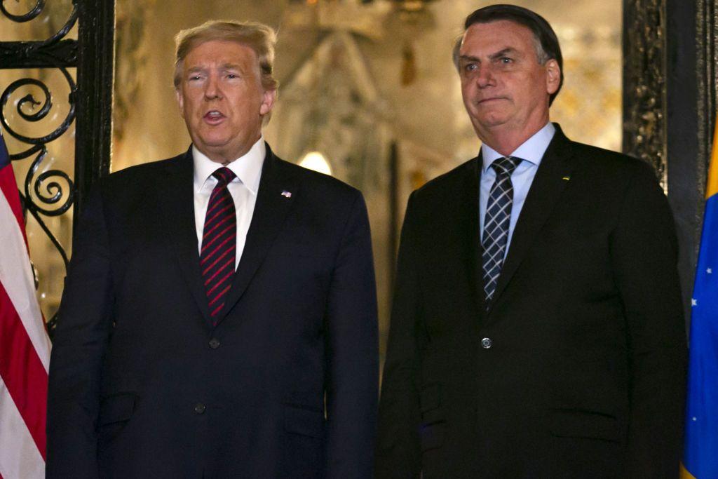 Donald Trump vistiendo una chaqueta oscura, camisa blanca y corbata roja está de pie junto a Jair Bolsonaro, quien viste una chaqueta oscura, una camisa azul con una corbata oscura con rayas claras.