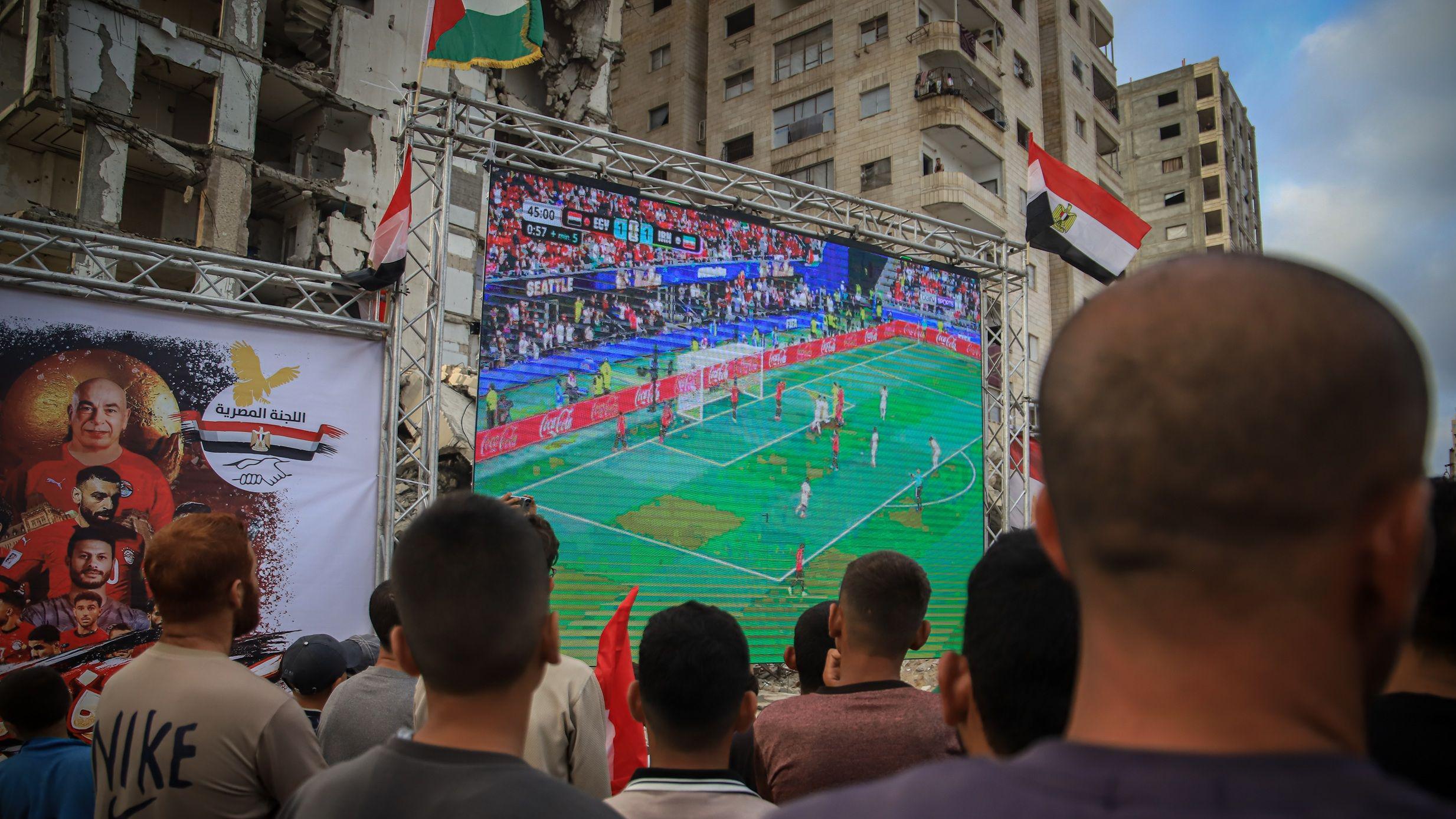تجمع فلسطينيون يحملون الأعلام لمشاهدة مباراة المجموعة السابعة من الدور الأول لكأس العالم لكرة القدم 2026 بين مصر وإيران على شاشة عملاقة نُصبت بين أنقاض المباني المدمرة، وذلك على الرغم من استمرار الهجمات الإسرائيلية على مدينة غزة في 27 يونيو/حزيران 2026.